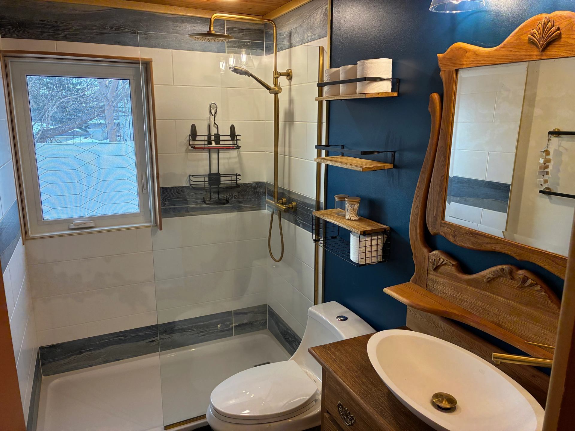 Bathroom with glass shower, brass fixtures, wooden vanity and shelves against blue wall.