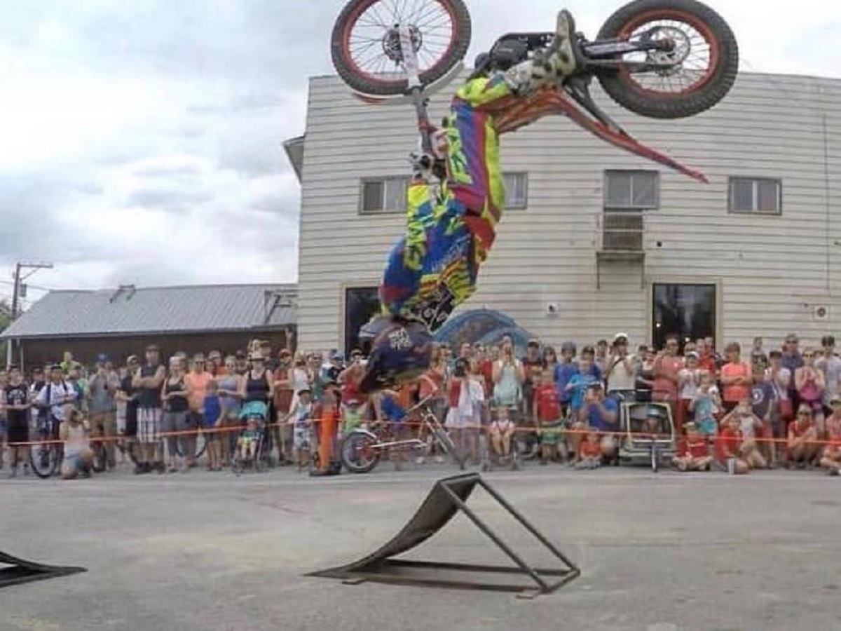 Motorcyclist in colourful gear performs upside-down jump off ramp before large outdoor crowd.