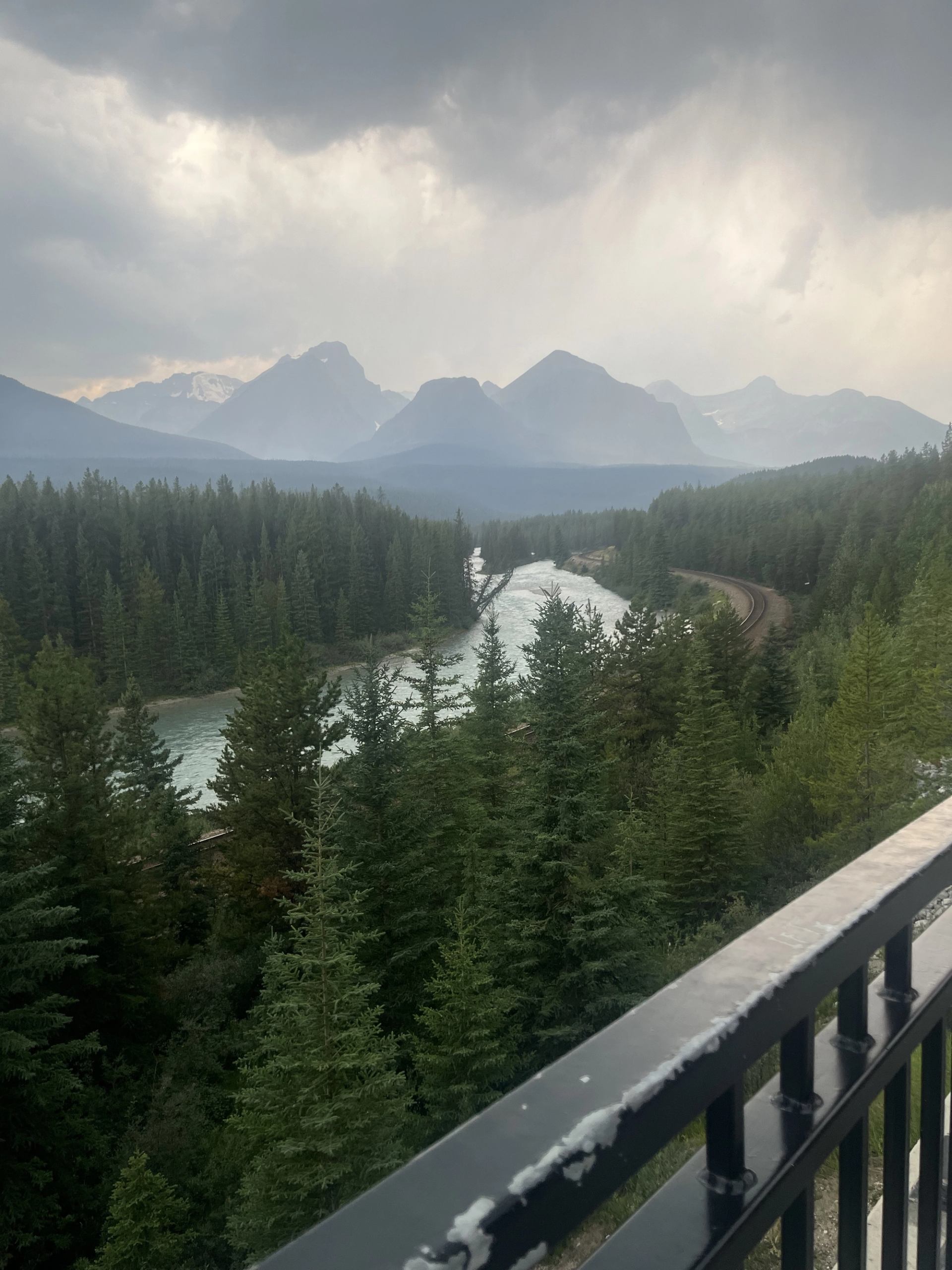 River and railway curve through dense evergreen forest below distant mountain range.
