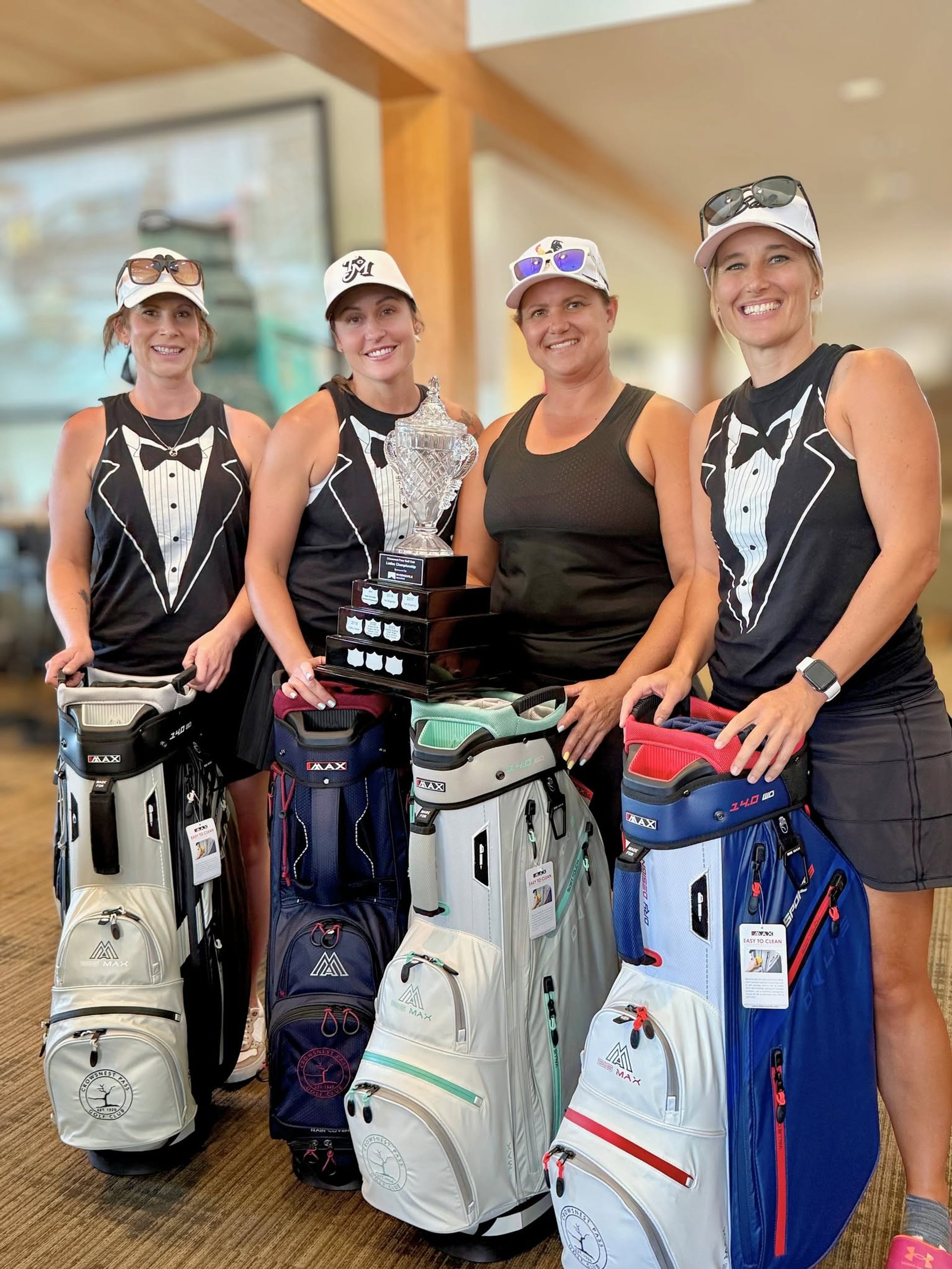 Four golfers in tuxedo shirts stand indoors with golf bags and a trophy.