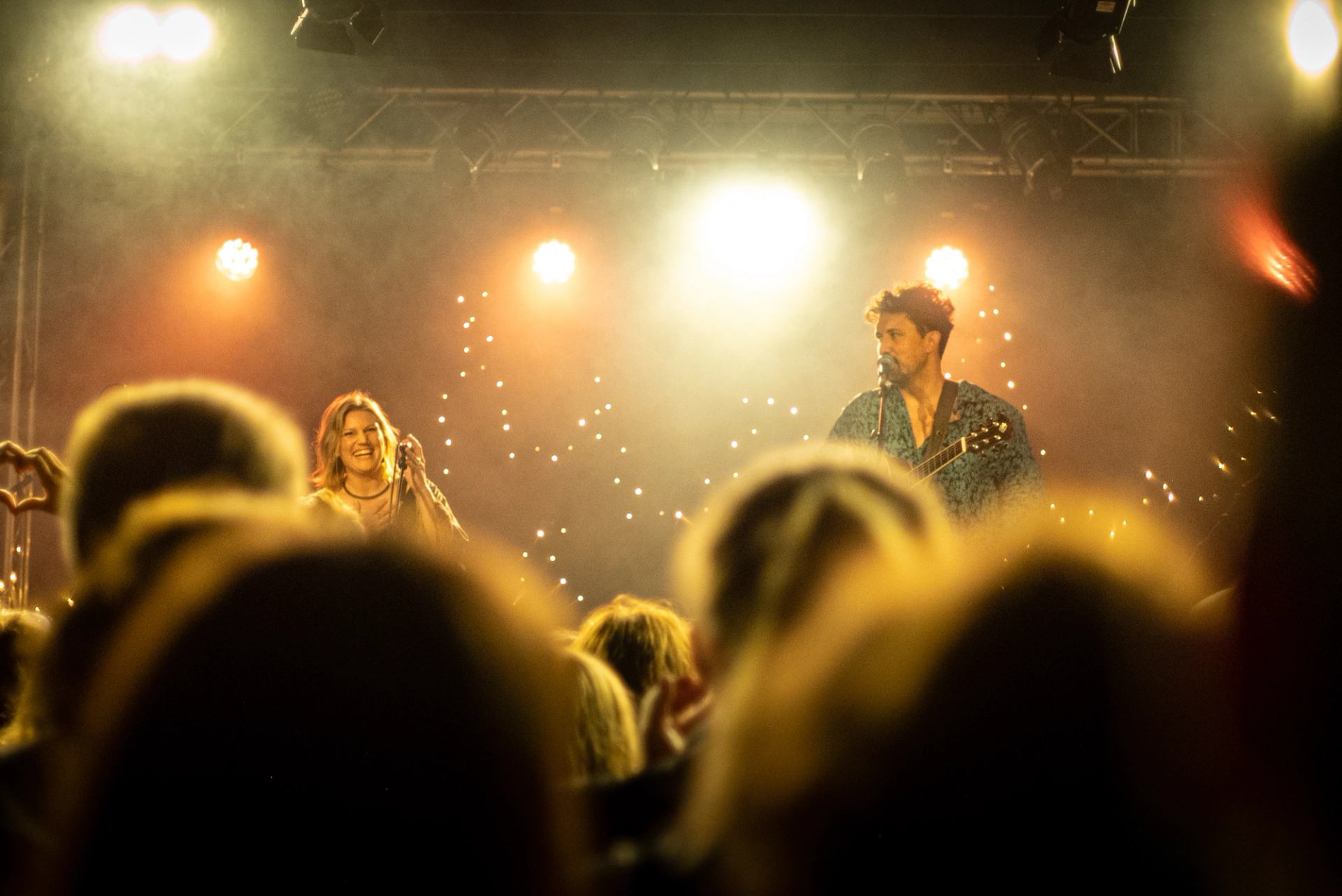 Musicians sing and play guitar on a brightly lit stage before a crowd.