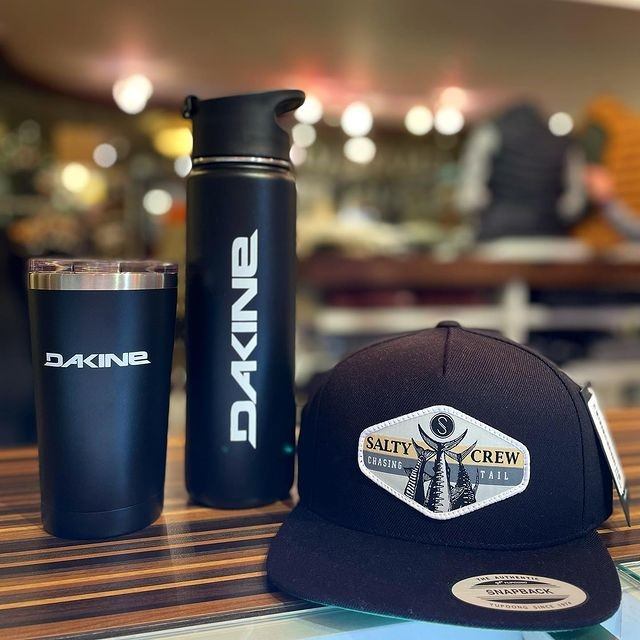 DAKINE tumbler, water bottle, and SANTA CRUZ cap on a wooden surface.