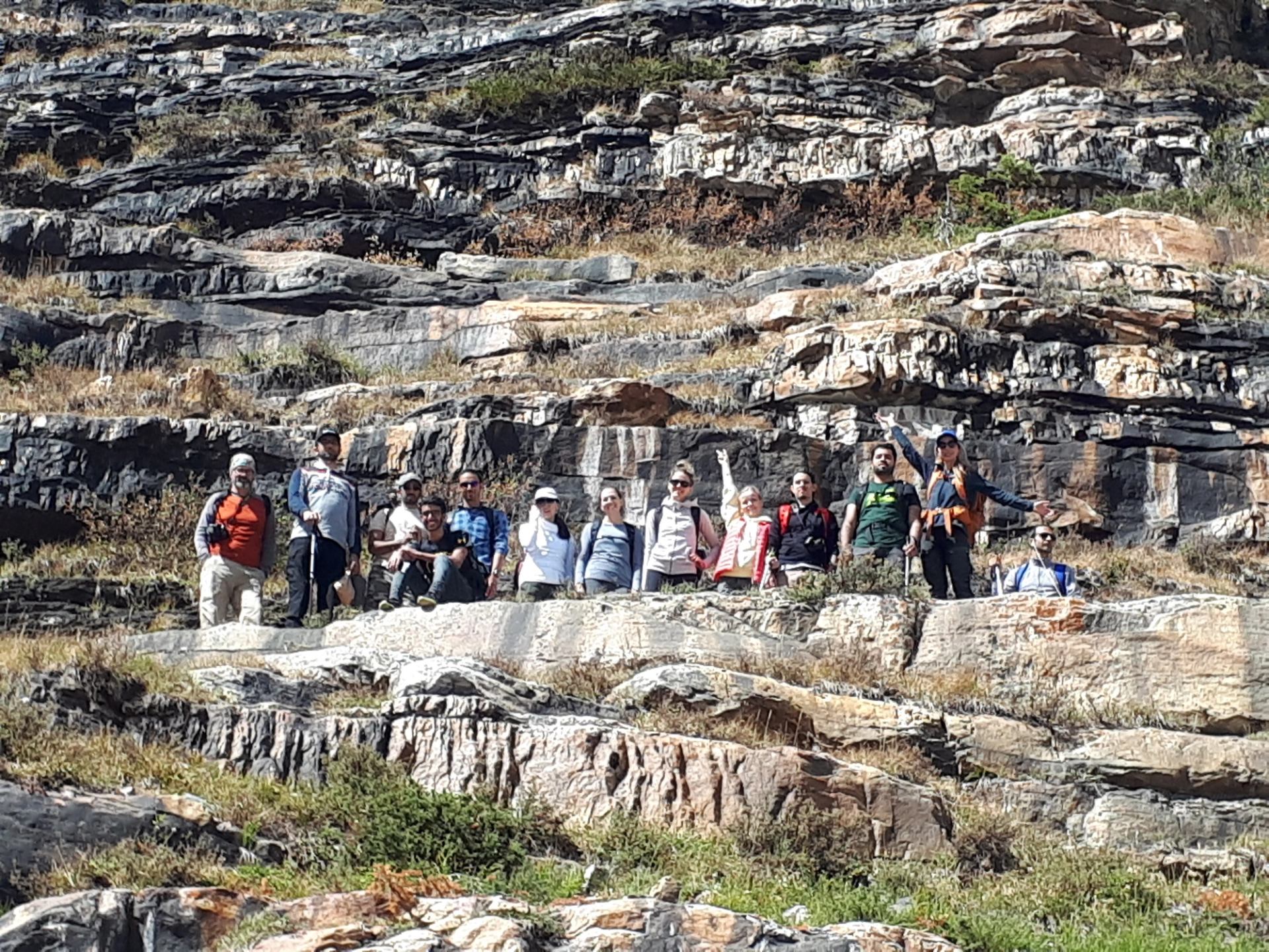 Hikers stand on layered rocky cliff, wearing backpacks and outdoor clothing.