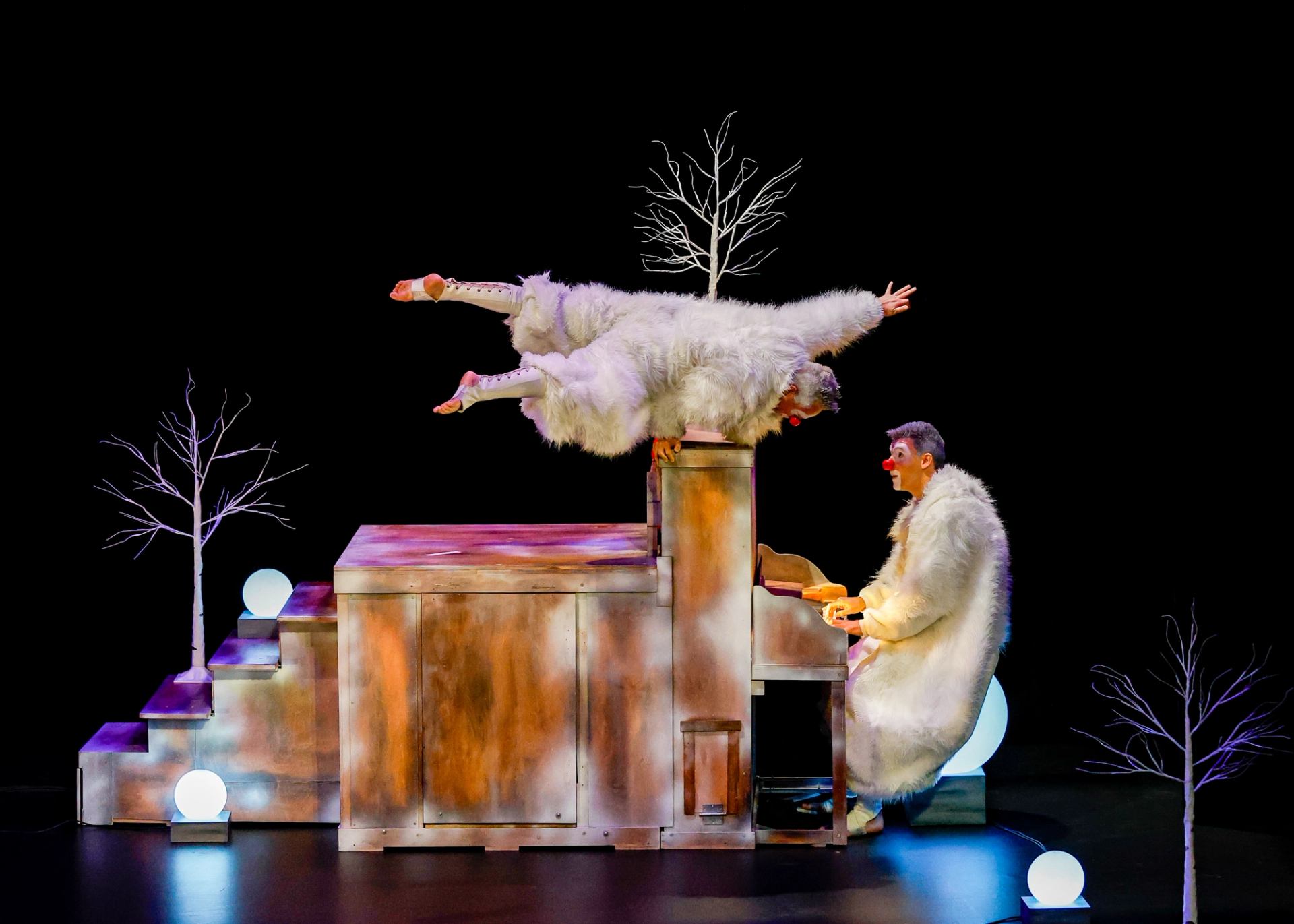 Stage clowns in white fur perform acrobatics on a piano amid glowing orbs.