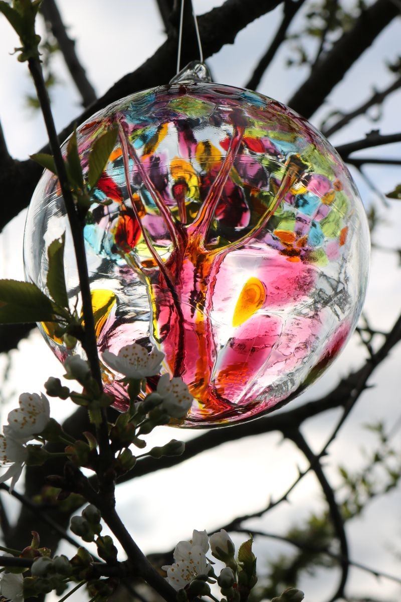 Colorful glass ornament hanging from tree branches with small white blossoms in background.