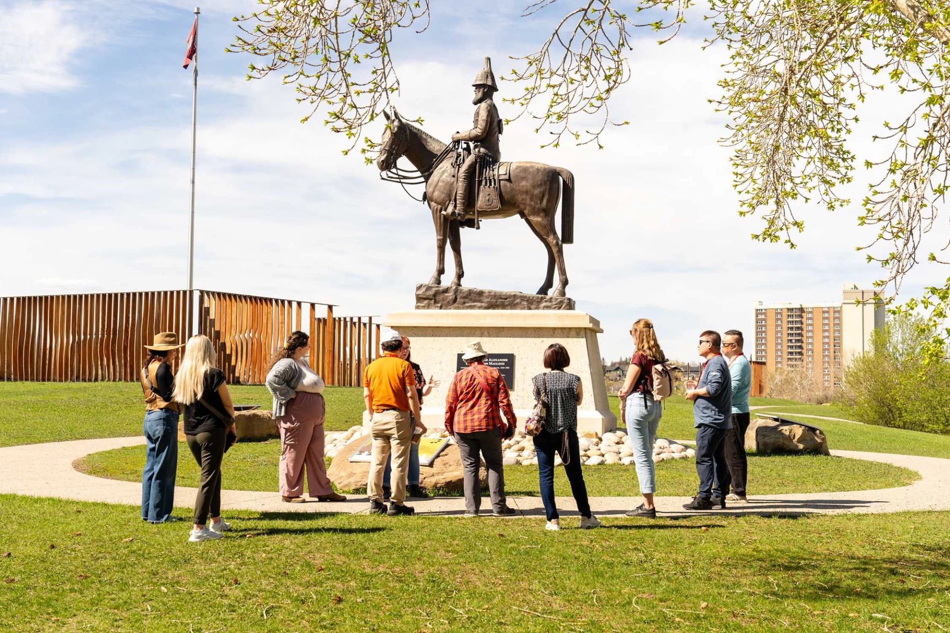 Group of people stand near equestrian statue in park with flagpole and buildings.