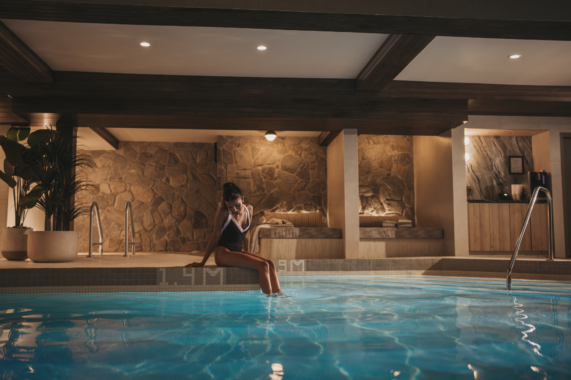 Woman sitting at the edge of an indoor pool, feet in water, in a spa room with stone walls and wood beams.