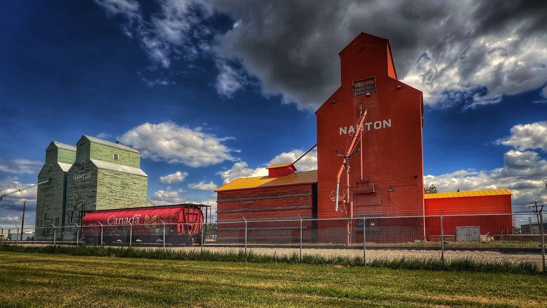 Green and red grain elevators with a Canada rail car under a cloudy sky.
