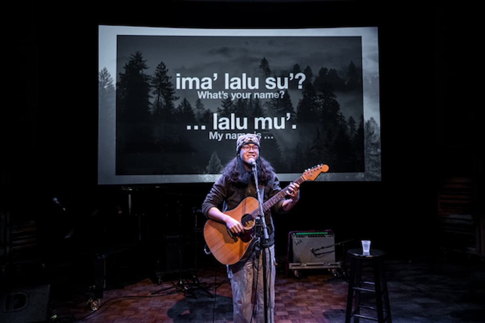 A musician performs with a guitar on stage in front of projected Indigenous language text and imagery.