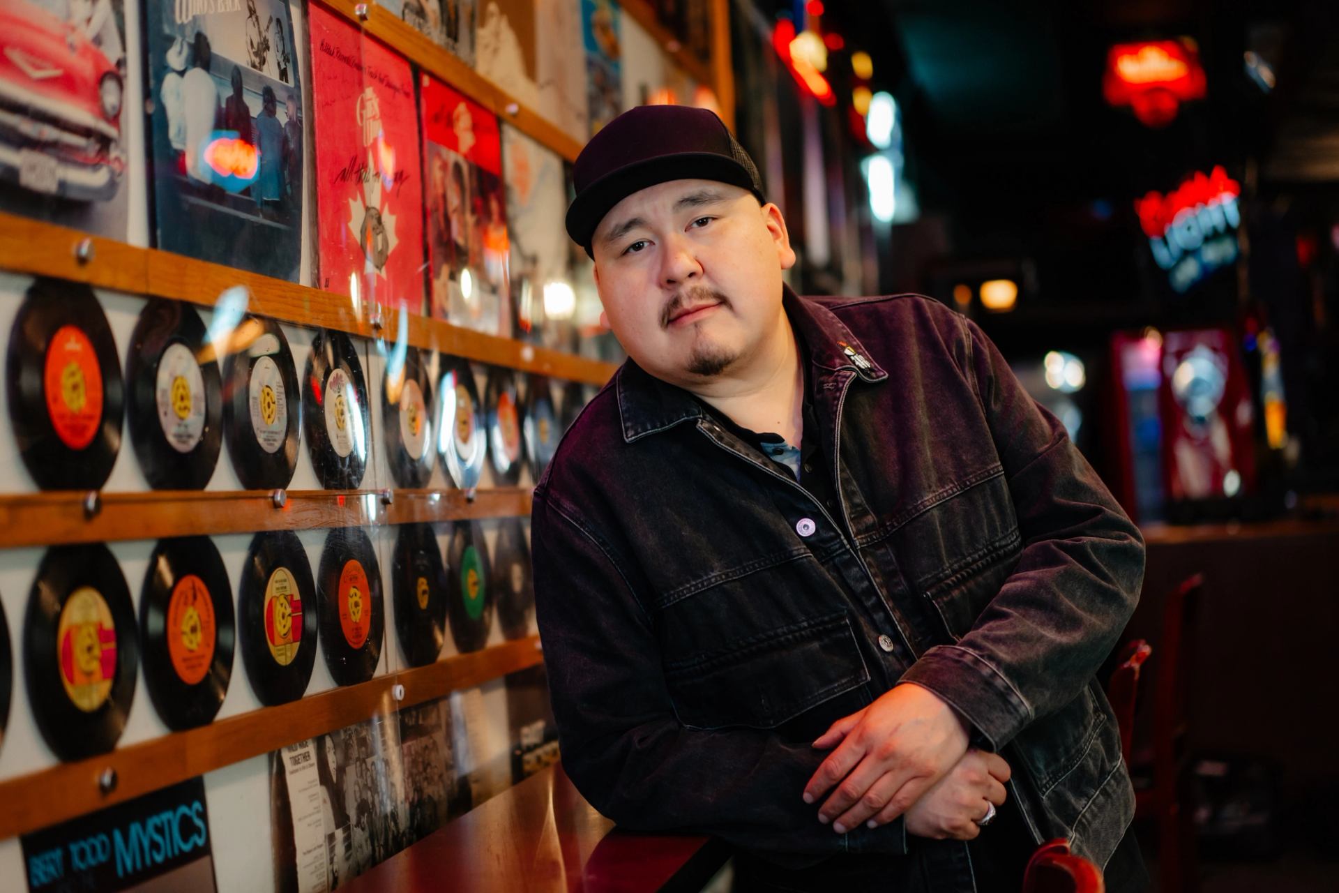 Person in dark jacket leans on bar beside wall of vinyl records and posters.