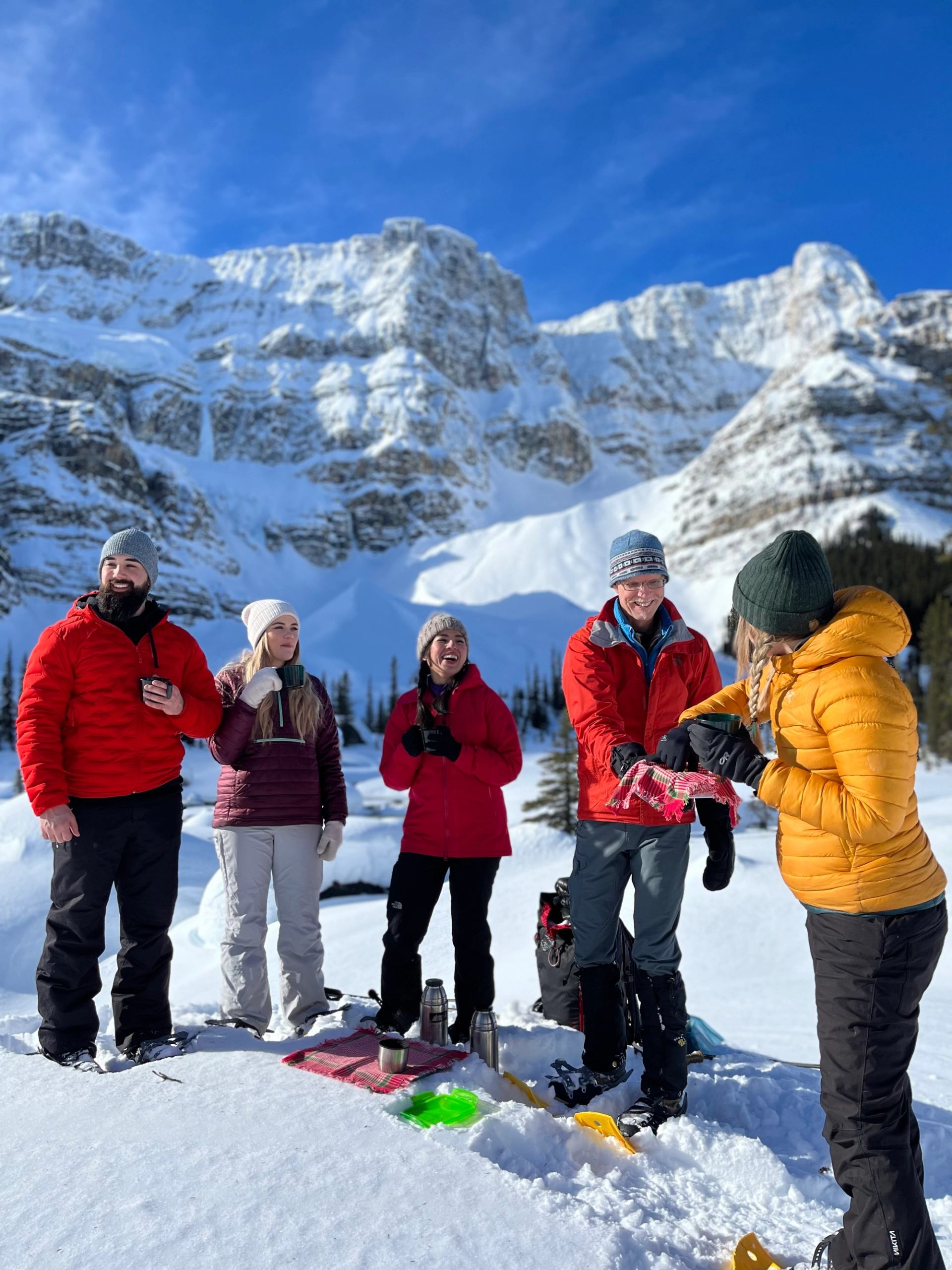Snowshoers enjoying hot chocolate and treats in front of mountains and glaciers.