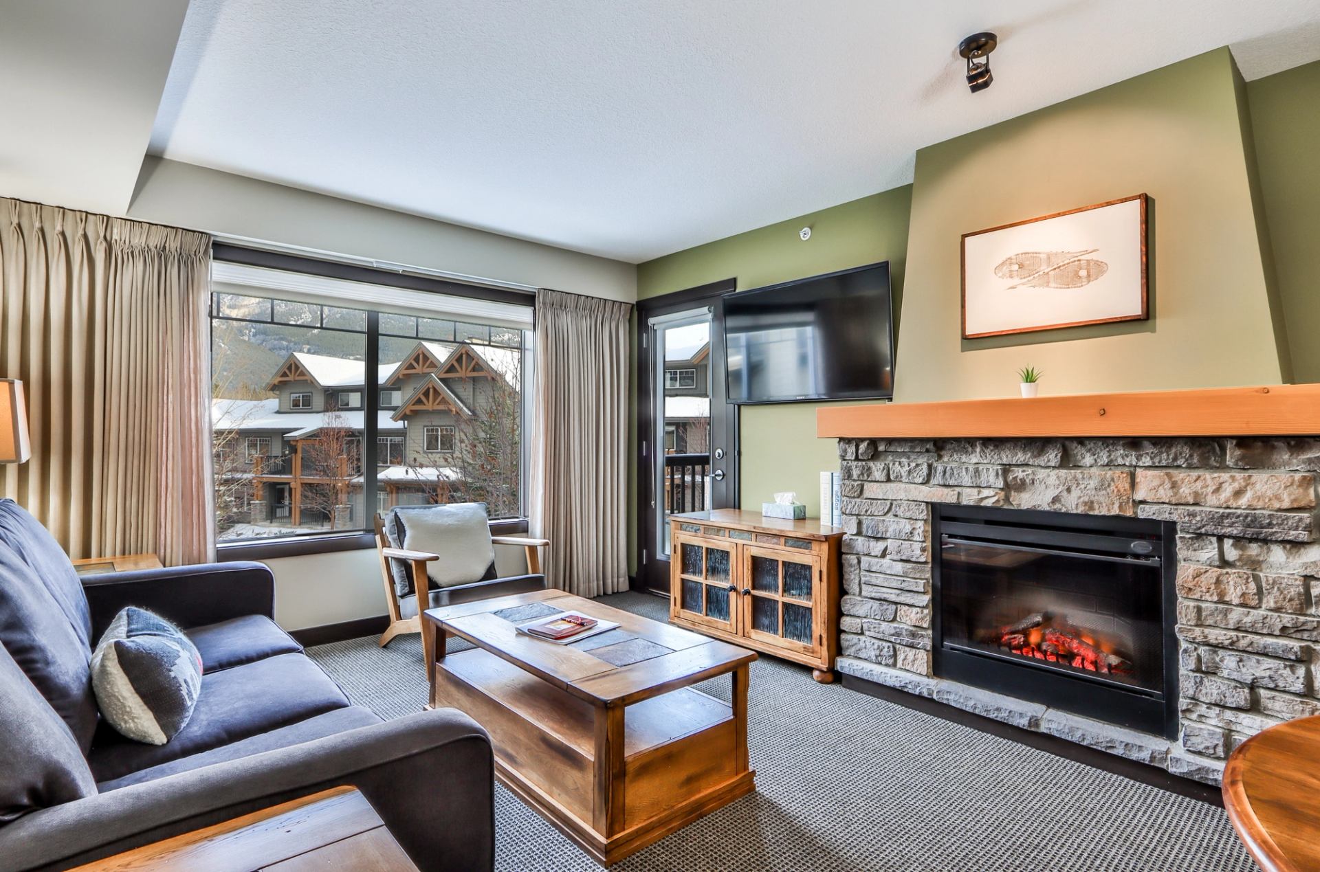 A cozy living room with a stone fireplace, a TV, a dark sofa, and a large window overlooking mountains.