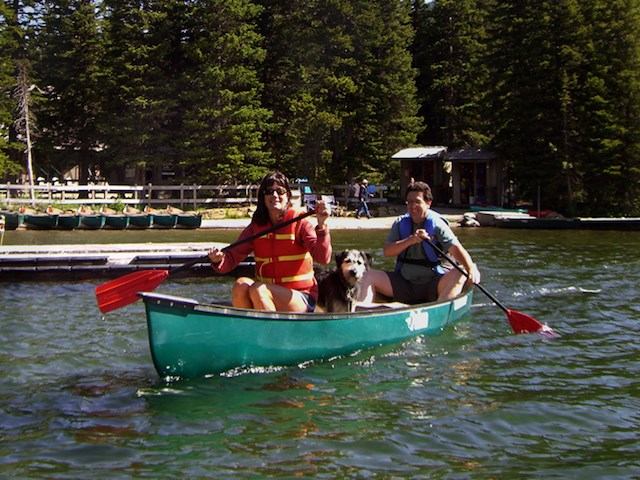 Two people and a dog paddling a green canoe near a dock on a forested lake.