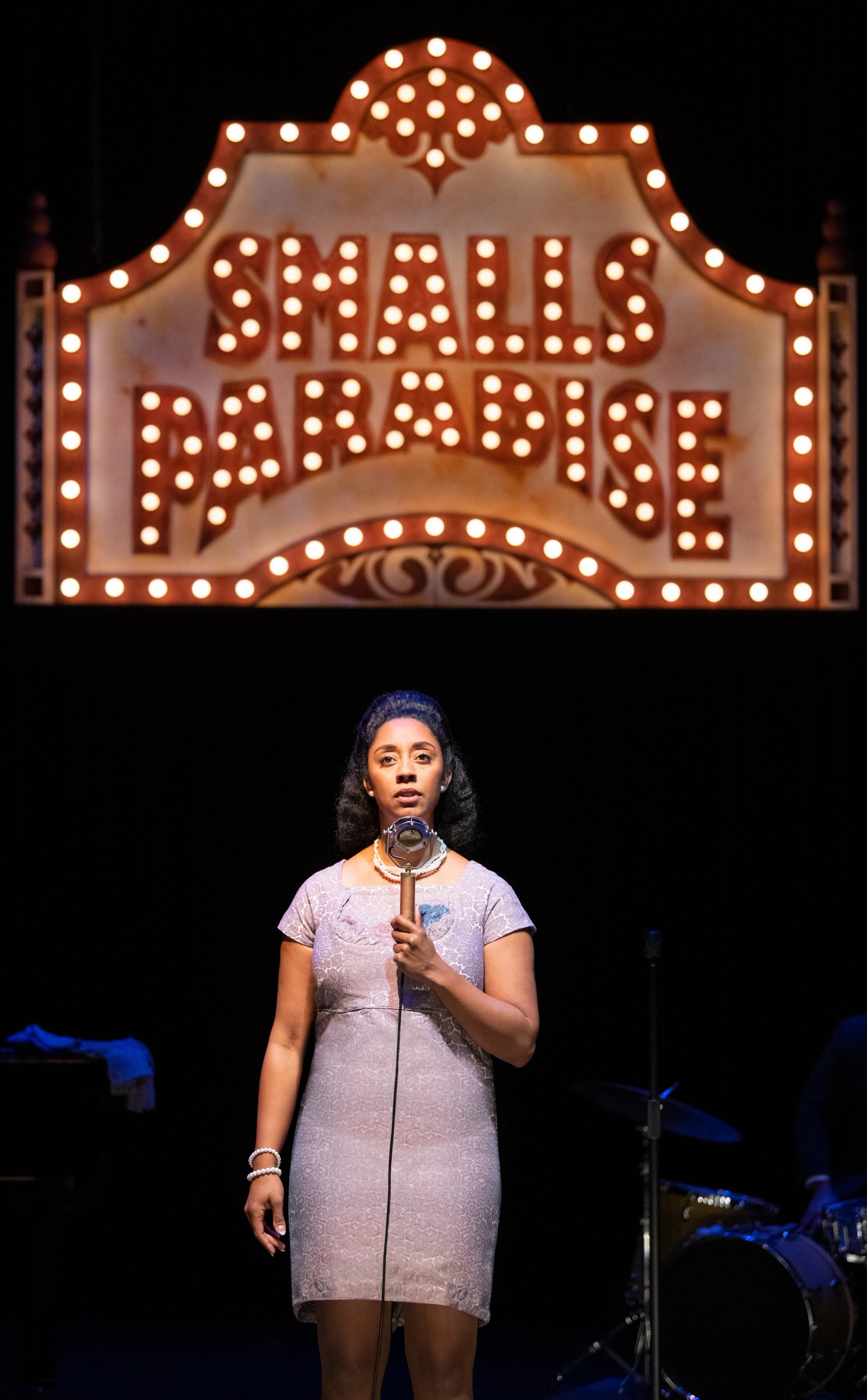 Singer in vintage dress stands at microphone under lit Smalls Paradise stage sign.