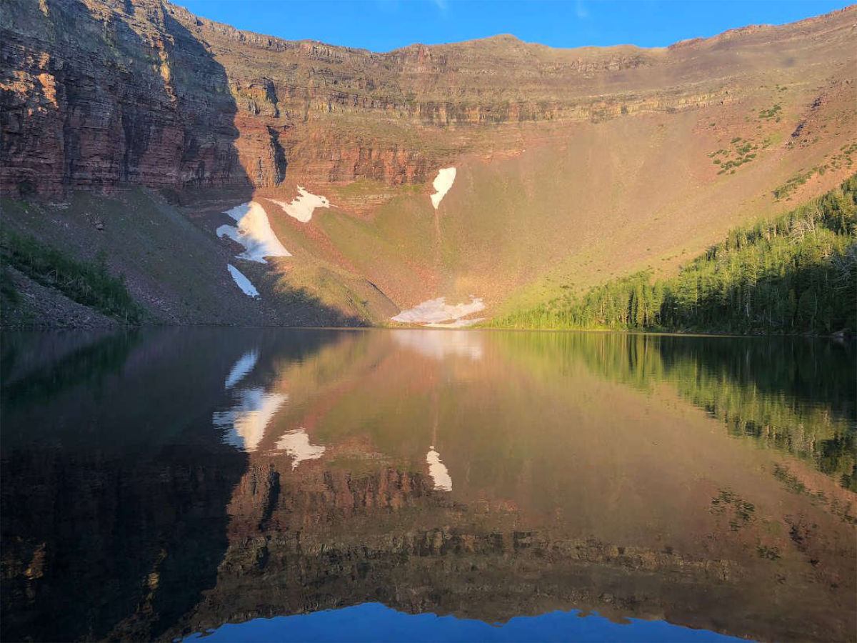 Mountain cirque with snow patches reflected in a calm alpine lake under clear sky.