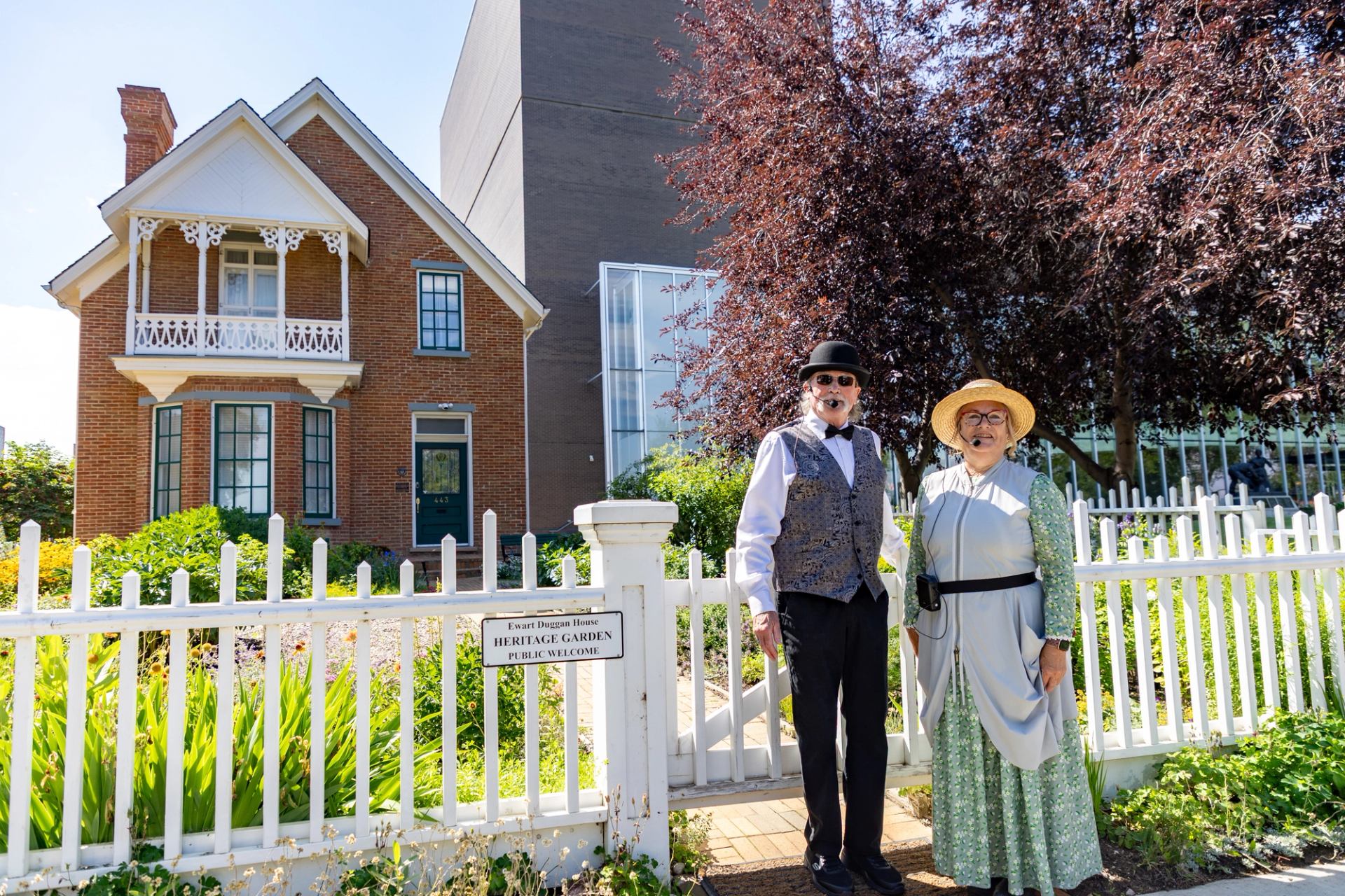 Two people in period clothing stand by white fence outside brick heritage house.