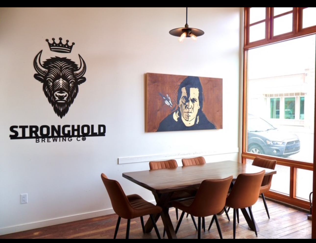 Interior of Stronghold Brewing Co. with a long table, chairs, wall logo, artwork, and large front windows.