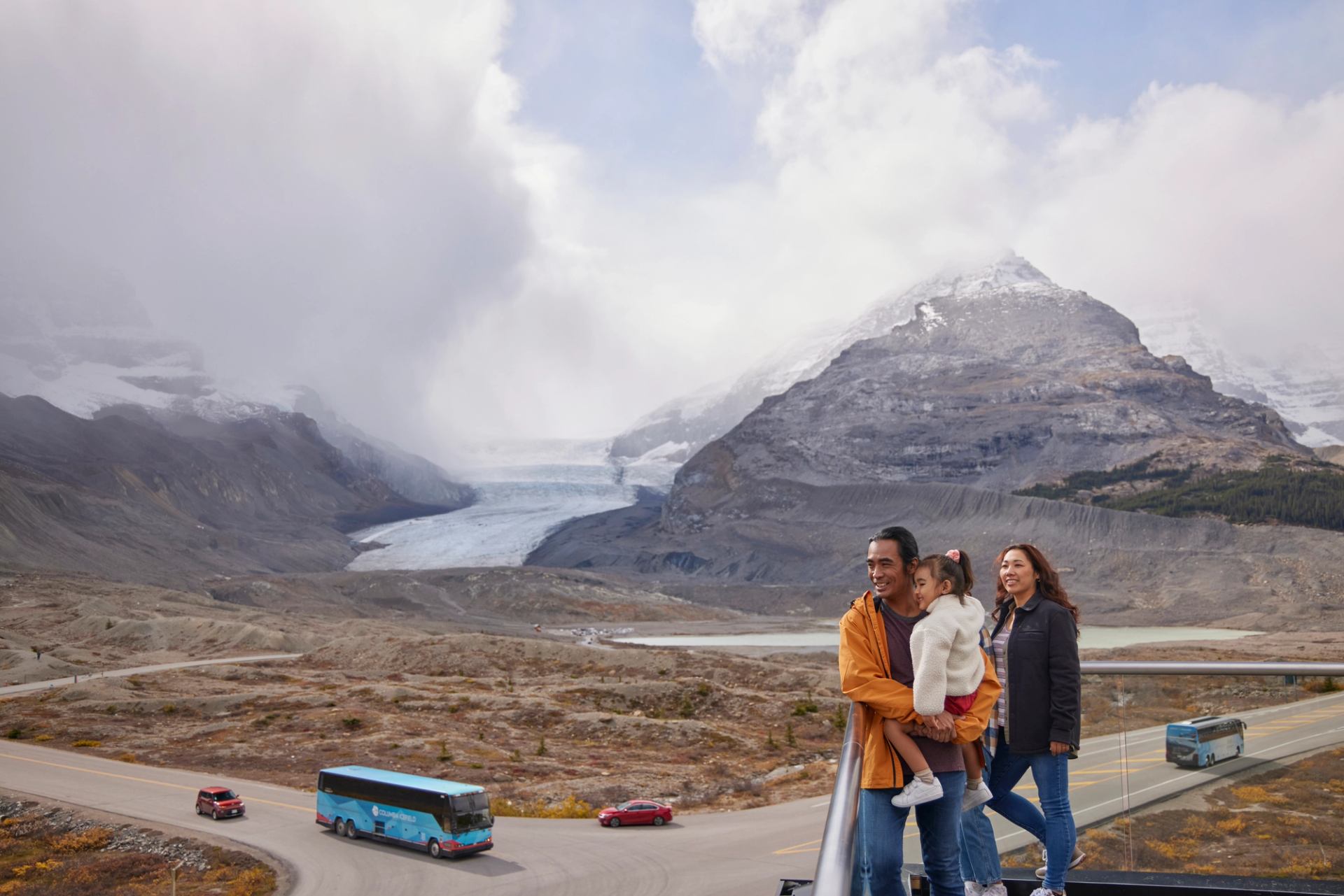 People standing at a high viewpoint overlooking a glacier, mountains, and tour buses.