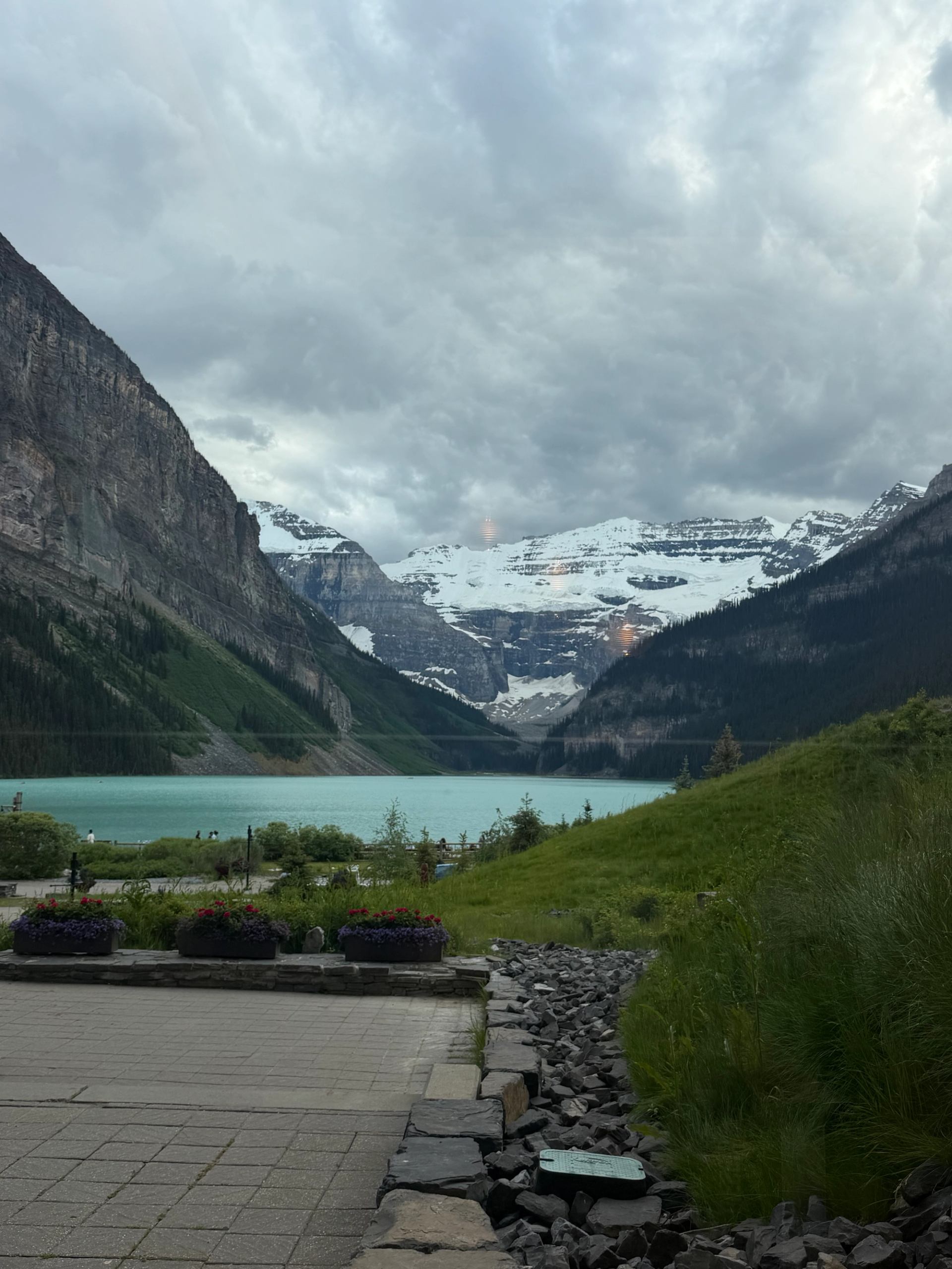 Turquoise lake below snow-covered mountains, with patio, flower planters and grassy slope.