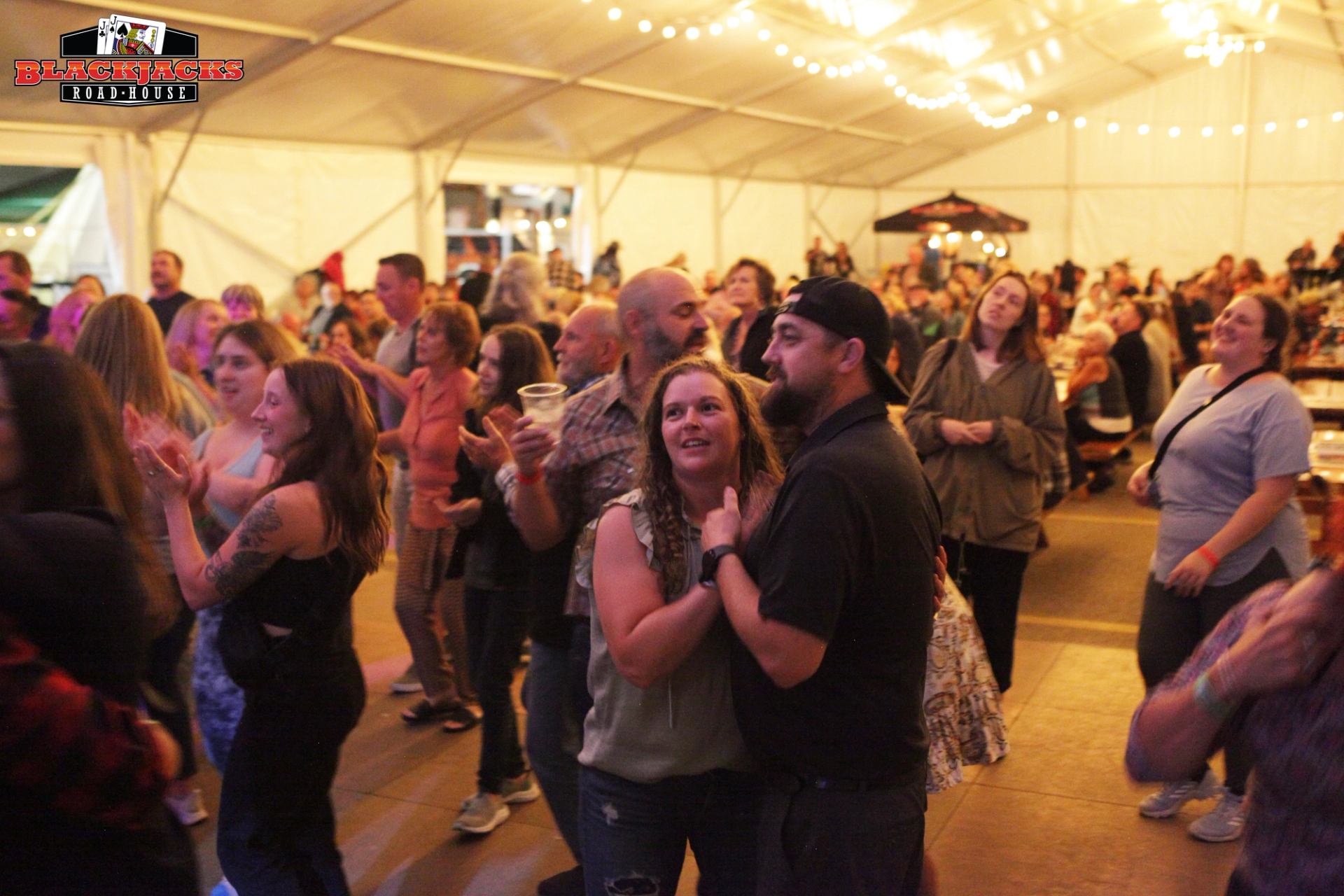 Crowd clapping at Blackjacks Road-House under string lights inside a tented concert hall.