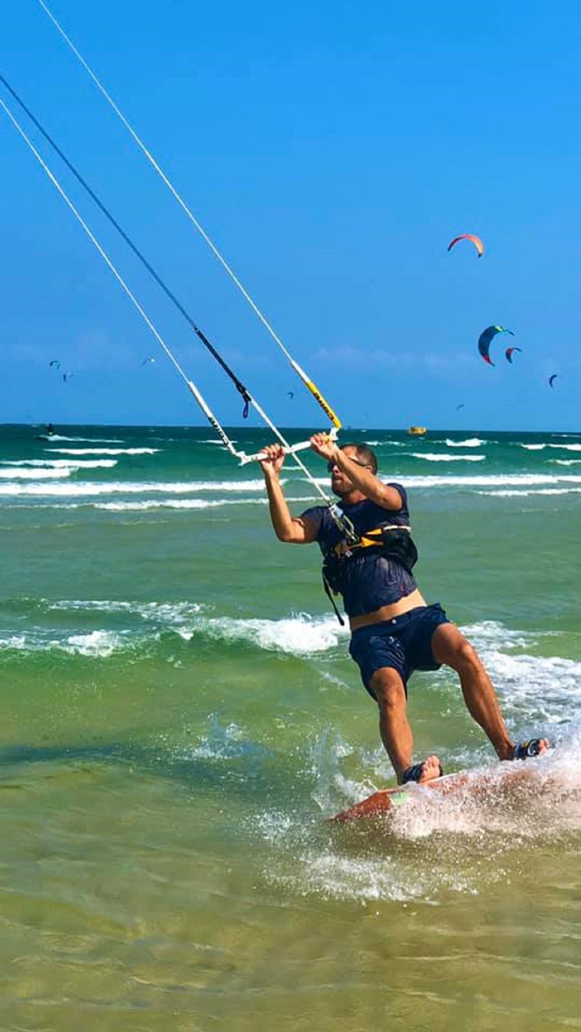 Kiteboarder carving through shallow ocean waves with multiple kites flying over turquoise water.
