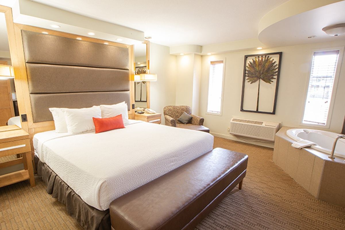 A hotel room showcases a large bed with white bedding, a padded headboard, and a bench at the foot. A seating area and windows bring in natural light, with a corner jetted tub visible across the room.