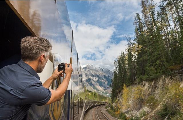 A passenger leans outside a train window to capture a photo of a mountain.