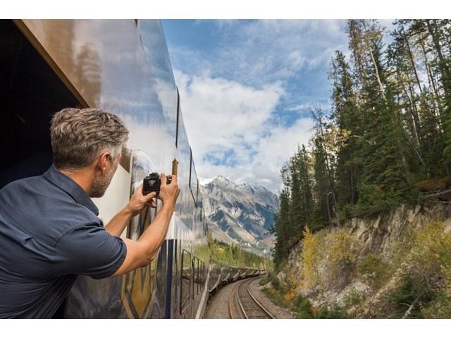 A passenger leans outside a train window to capture a photo of a mountain.
