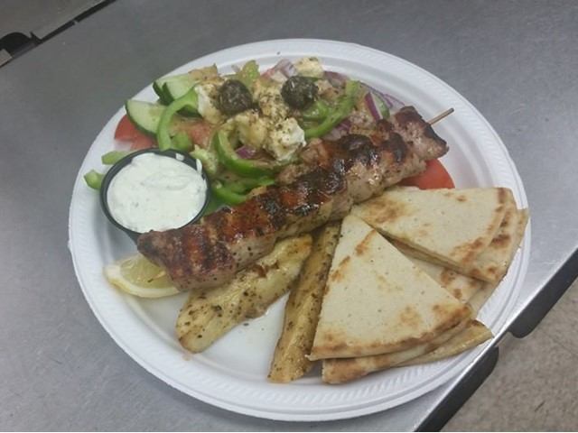 Greek plate with grilled skewer, pita wedges, salad, potato wedges, and tzatziki.