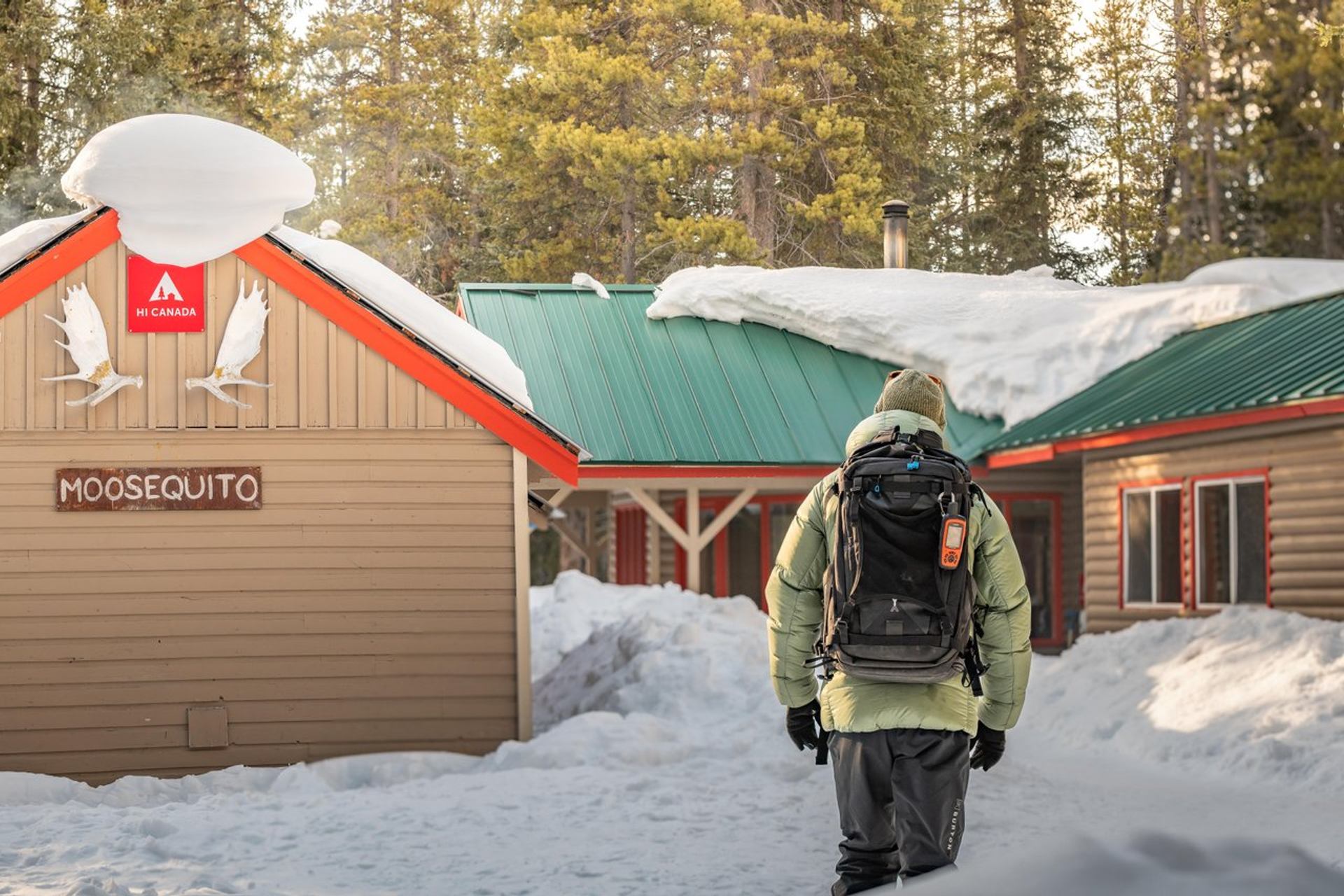 Winter exterior of HI Mosquito Creek with snow-covered cabin buildings, traveller with backpack.