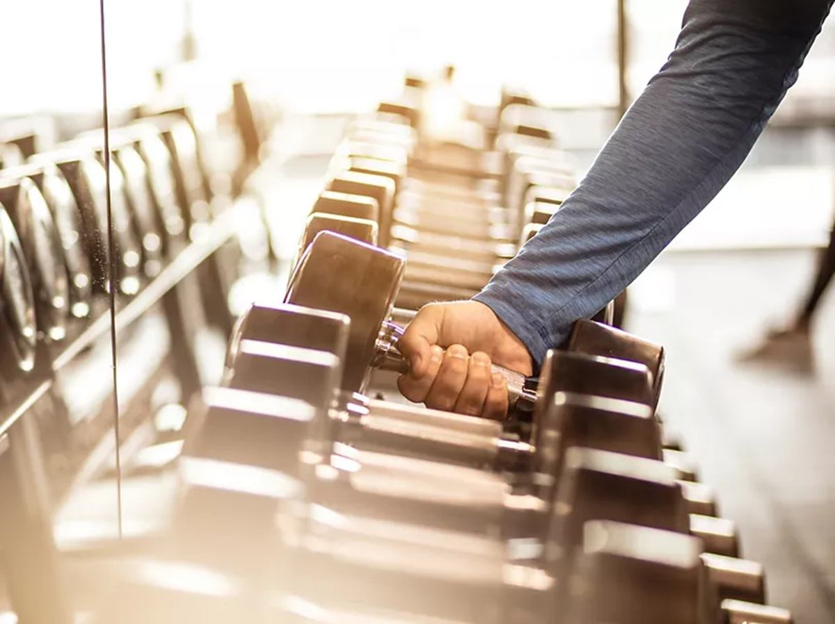 Arm in blue sleeve lifting a dumbbell from a rack in a gym.