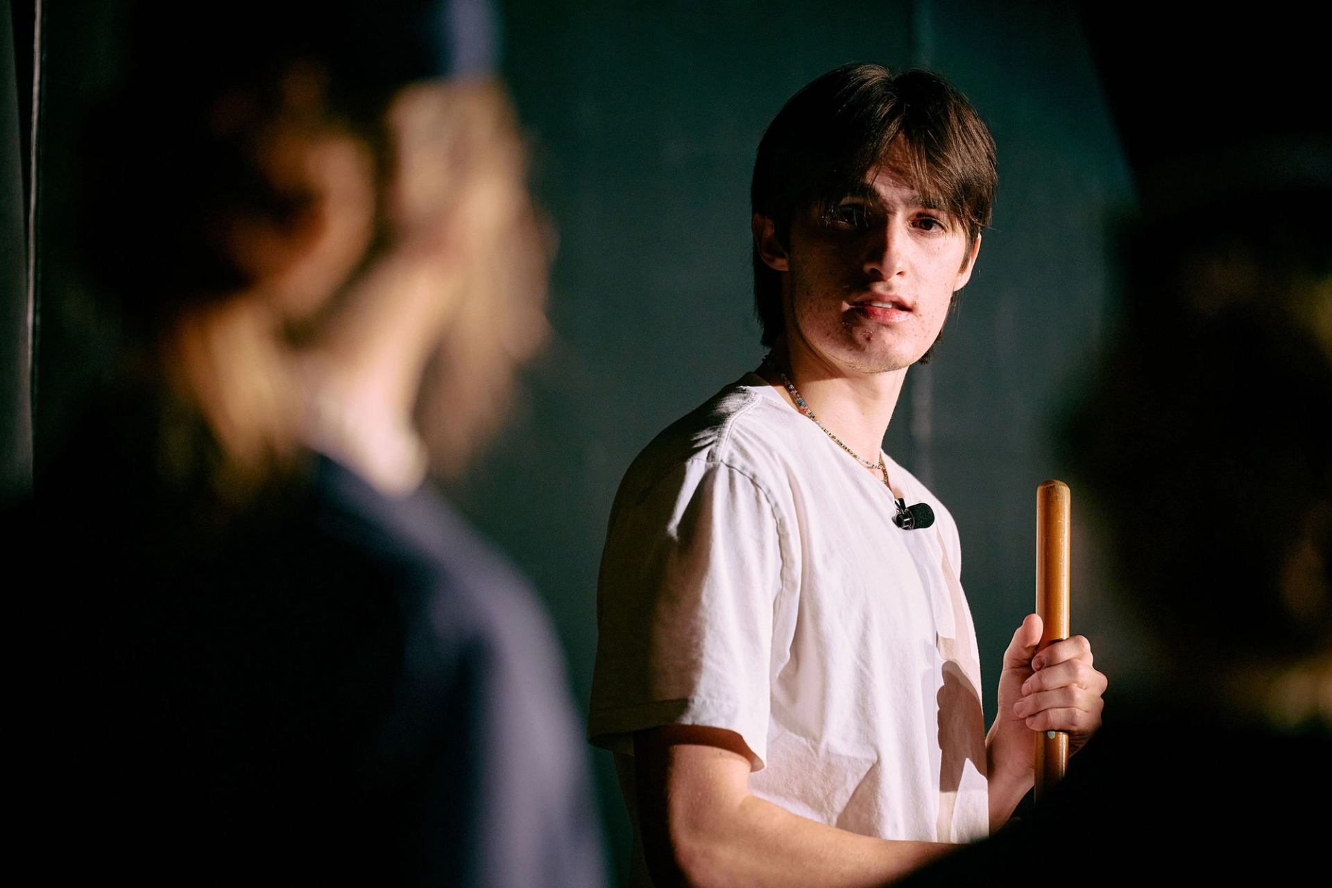 Stage performer in white shirt holds wooden prop, facing blurred figures under spotlight.