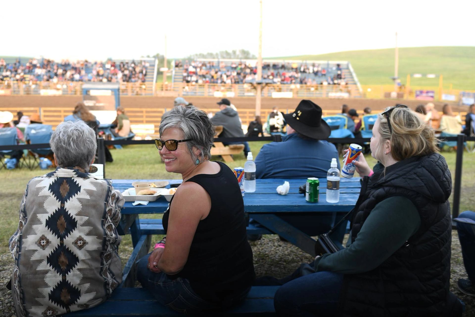 Visitors sit at picnic tables watching a rodeo event in an outdoor arena filled with spectators.
