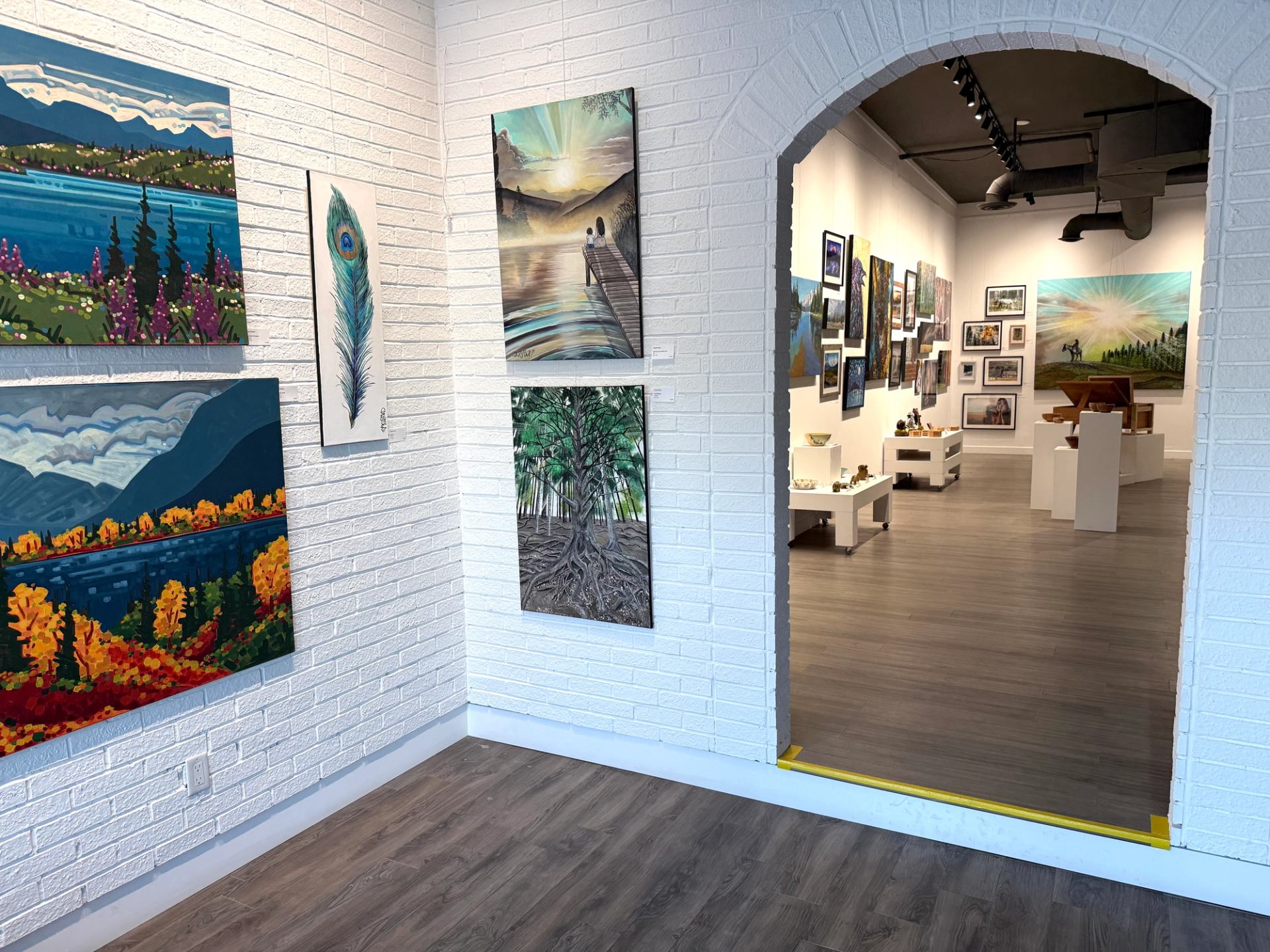 Art gallery interior with landscape paintings on white brick walls and wood flooring.