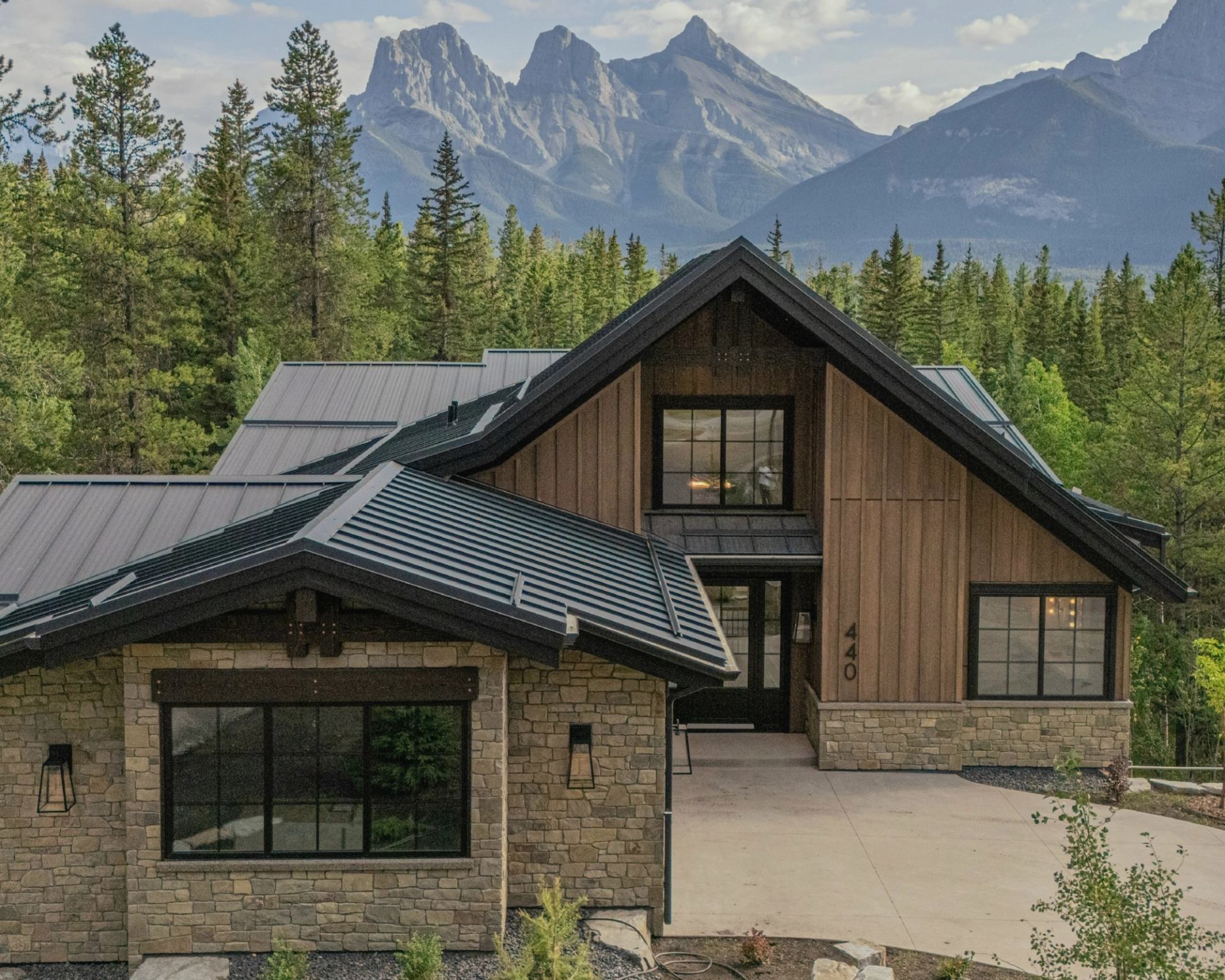 A modern stone-and-wood mountain home sits among evergreen trees with towering mountain peaks in the background.