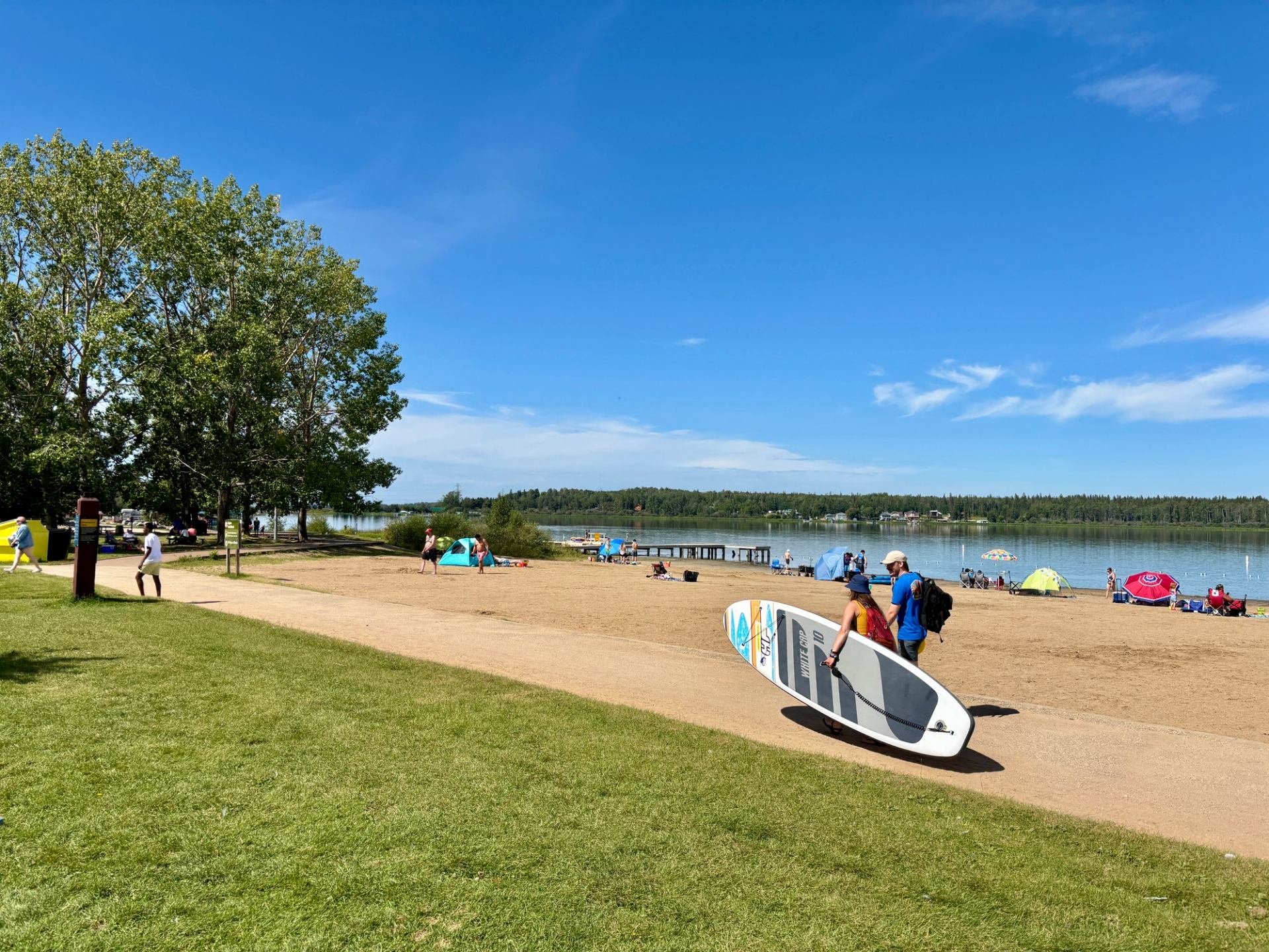 People walk with a paddleboard on sandy lakeside beach with tents and trees.