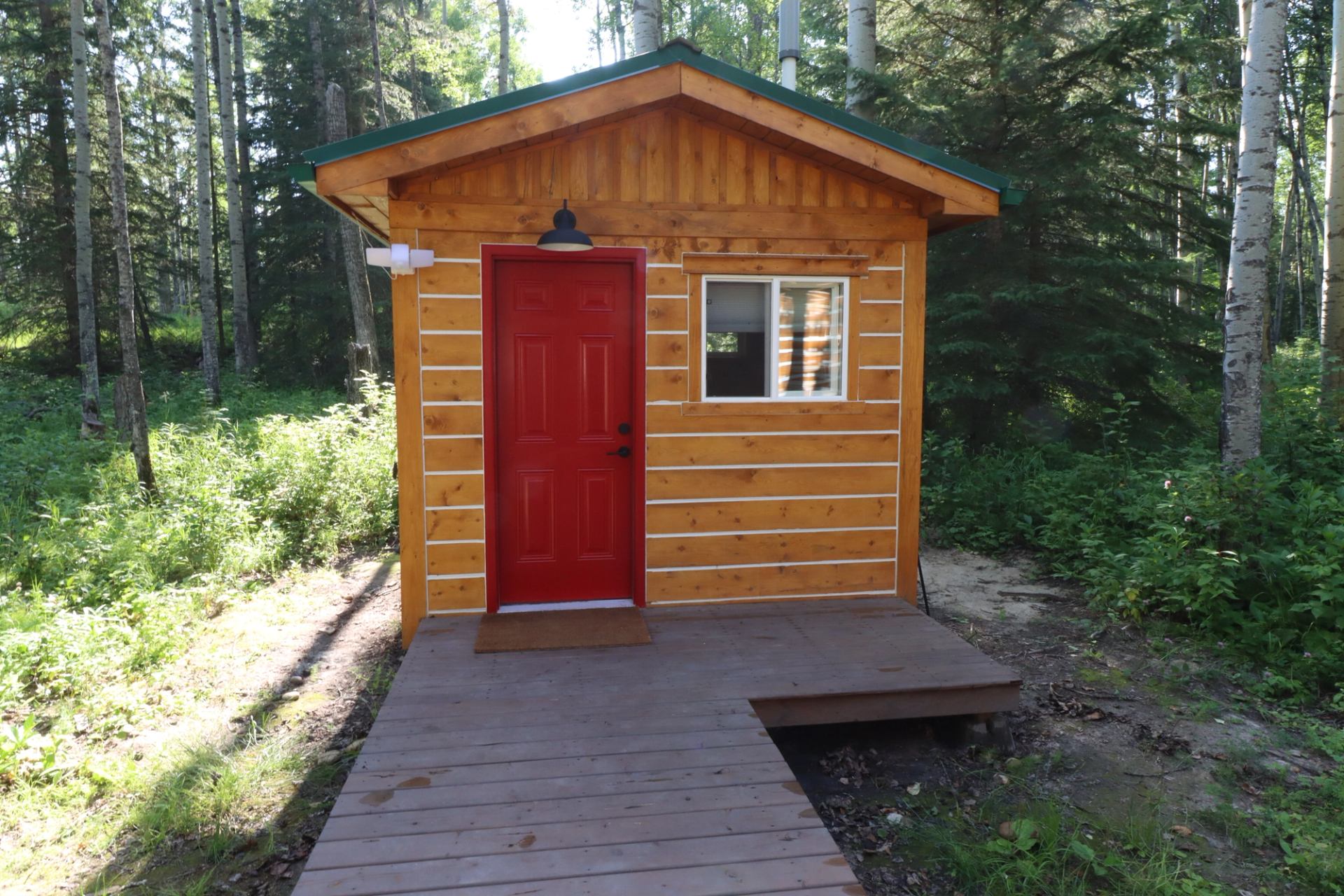 Small forest cabin with red door, window, and wooden walkway surrounded by trees and greenery.