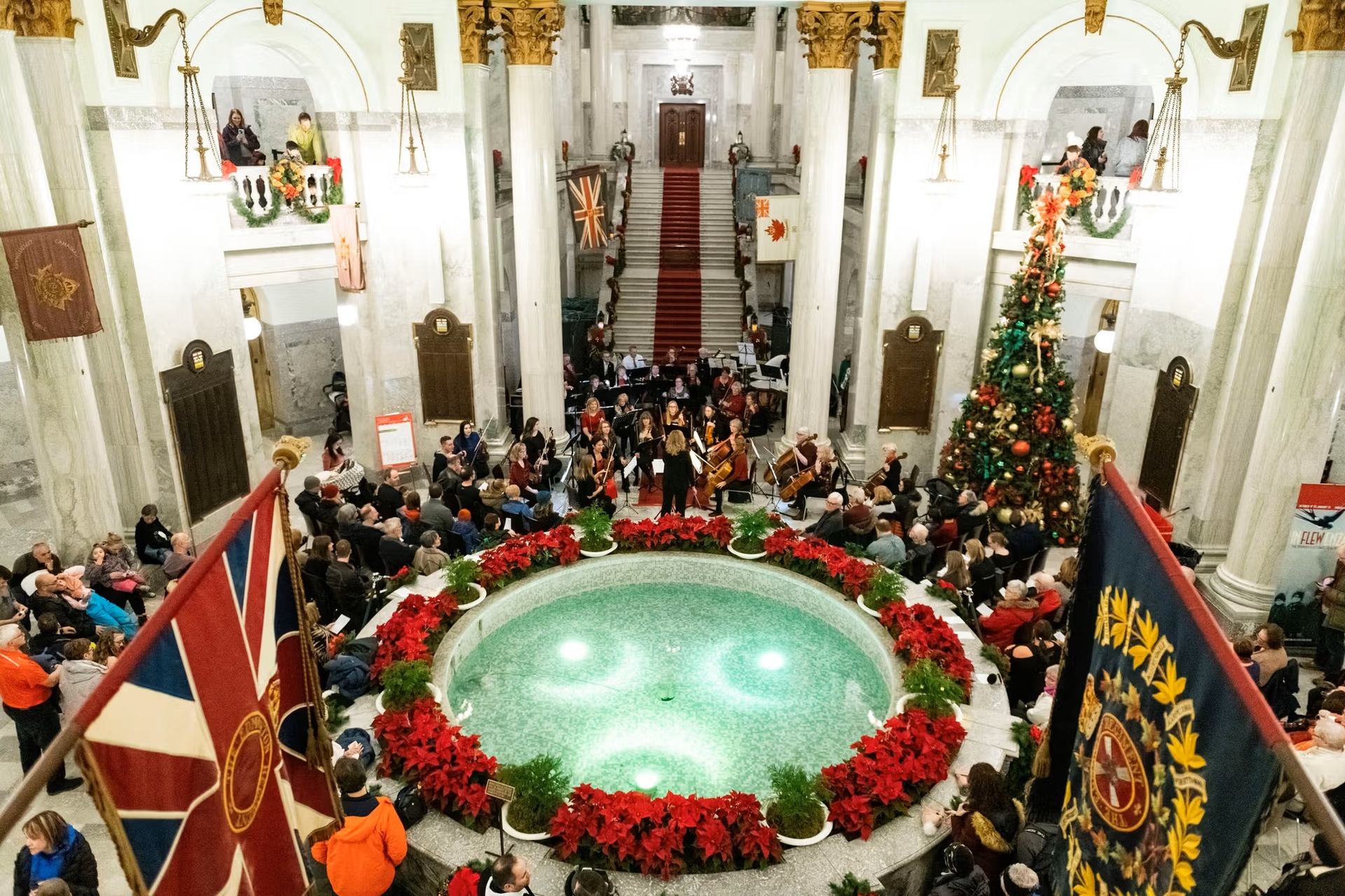 Crowd gathered around a fountain with poinsettias and Christmas tree inside Alberta Legislature.