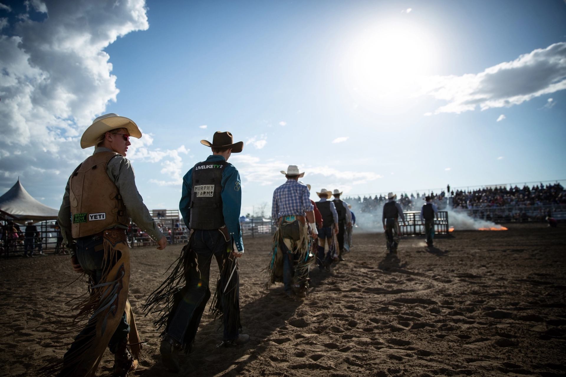 Rodeo riders in cowboy hats walk across dirt arena toward smoke and grandstand.