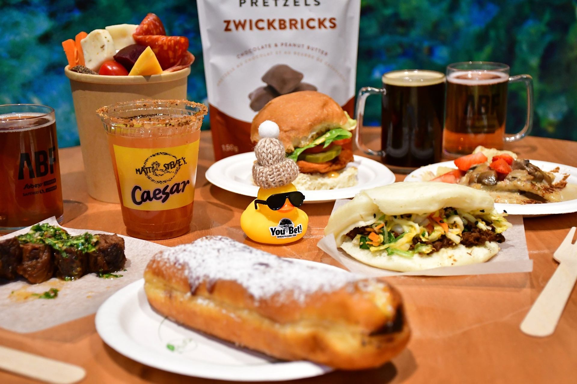 Assorted festival foods and drinks on a table, including burger, bao, pastries and beer.