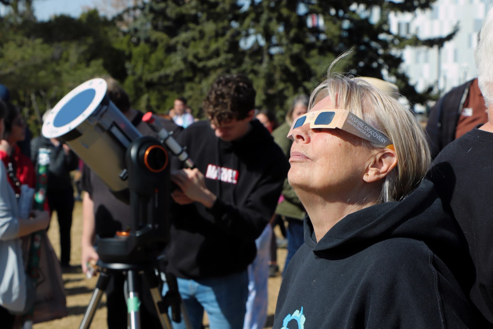 Person wearing eclipse glasses looks up near telescope at crowded outdoor event.
