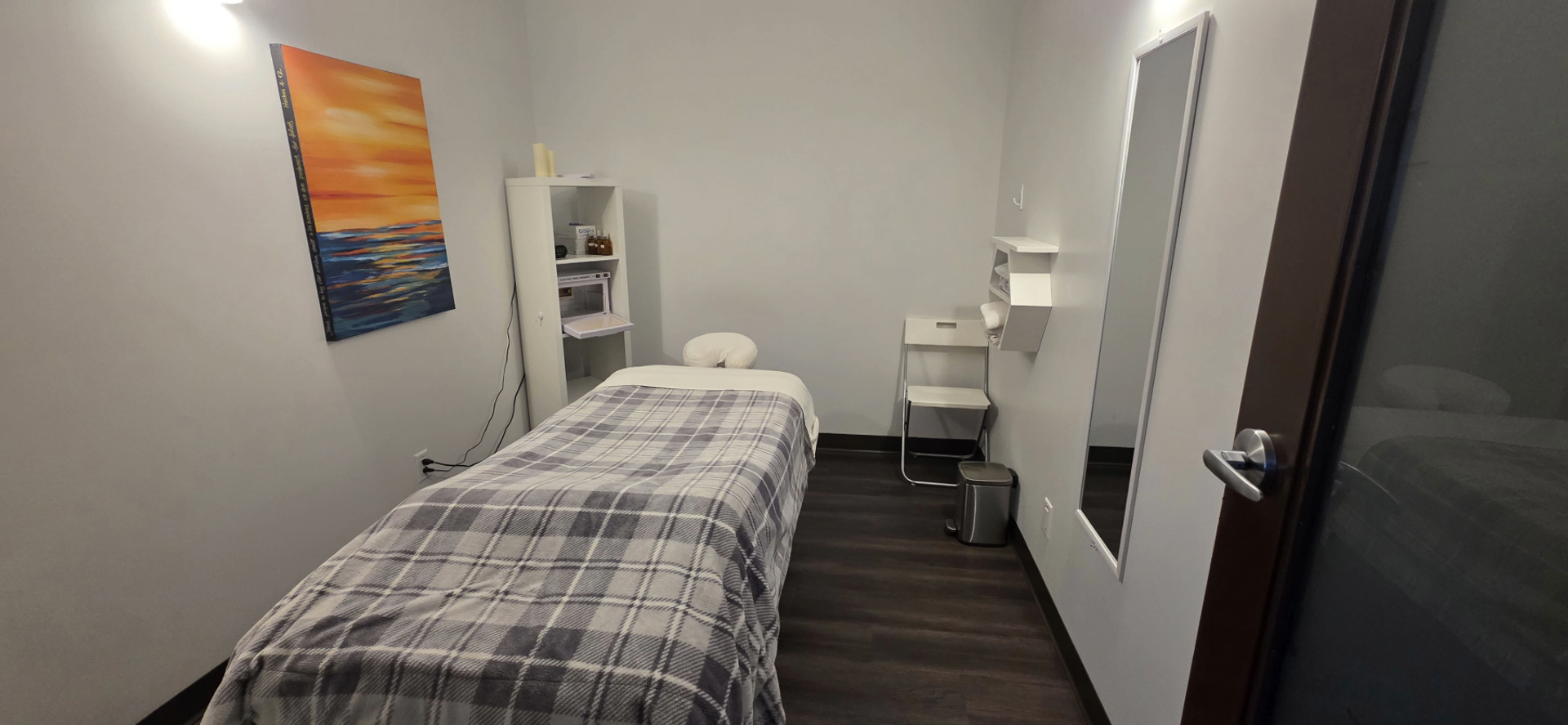 Treatment room with a covered table, shelving, chair, mirror and wall art.