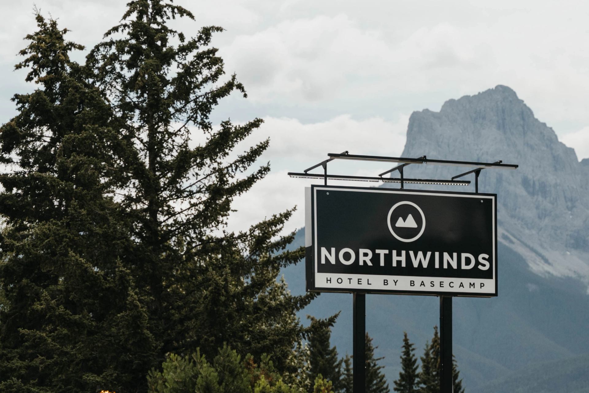 Northwinds Hotel by Basecamp roadside sign with evergreen trees and rocky mountain backdrop.