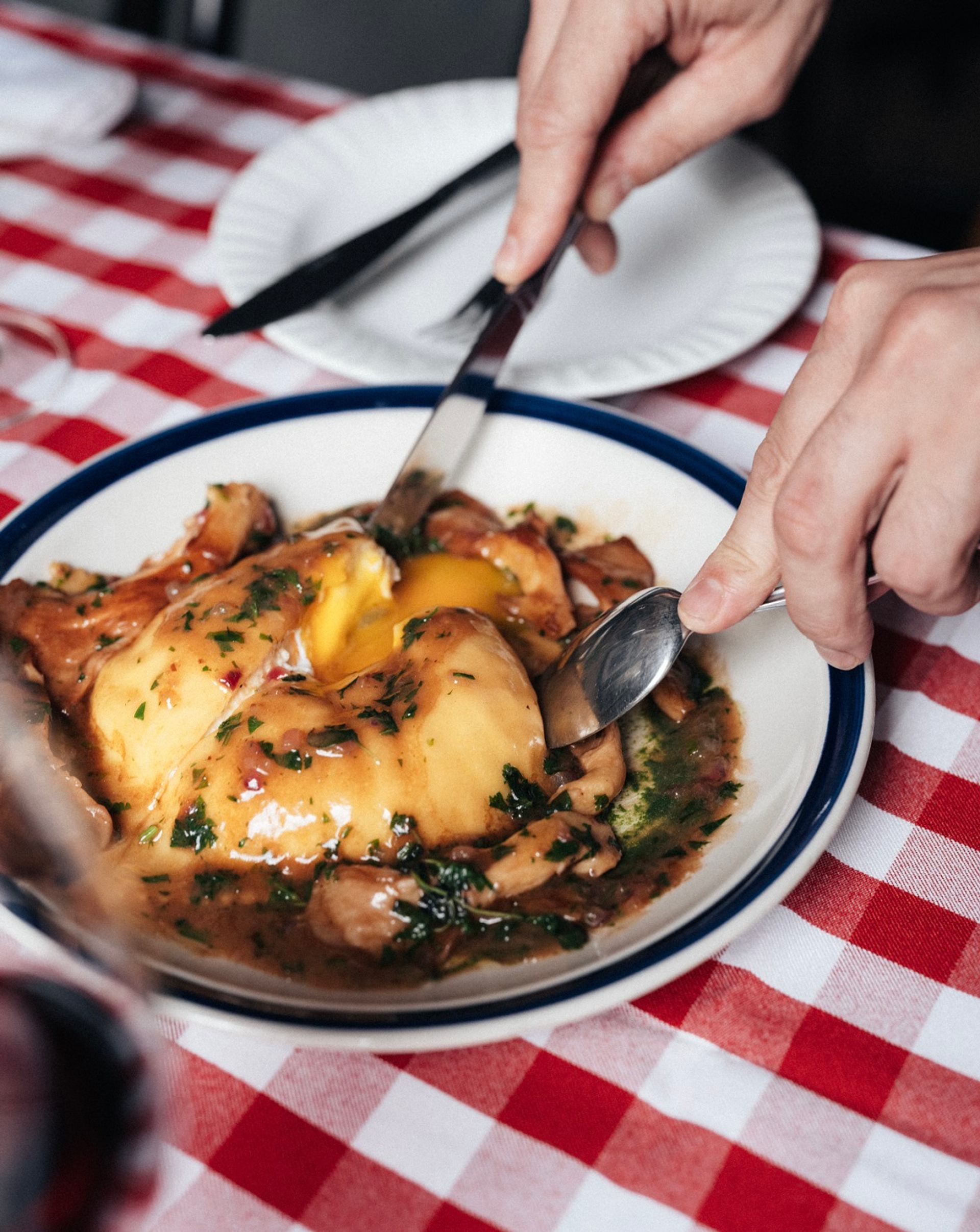 Hands cut into saucy chicken and egg dish on checkered tablecloth.