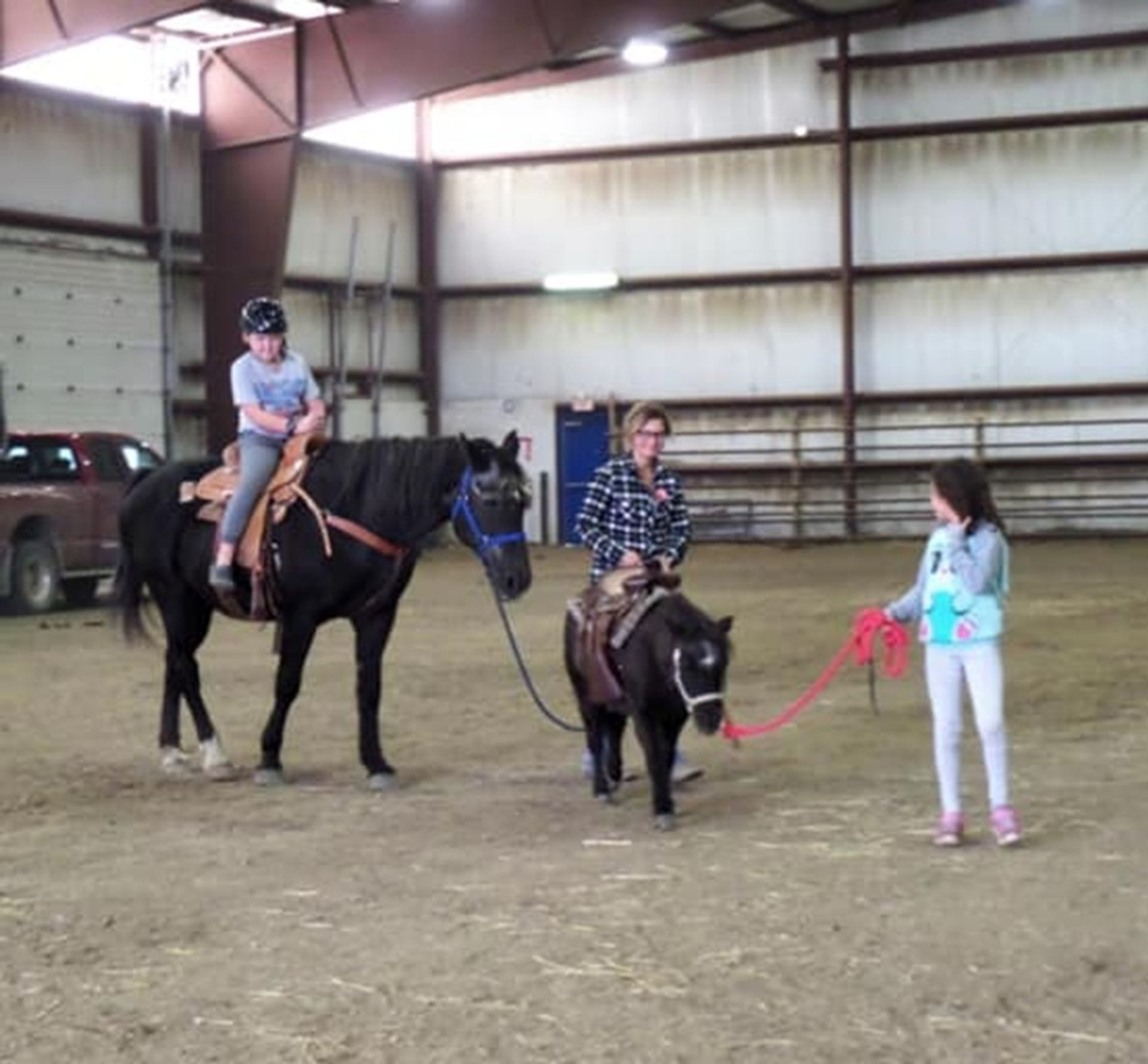 Indoor riding arena with child on horse and two people leading a small pony.