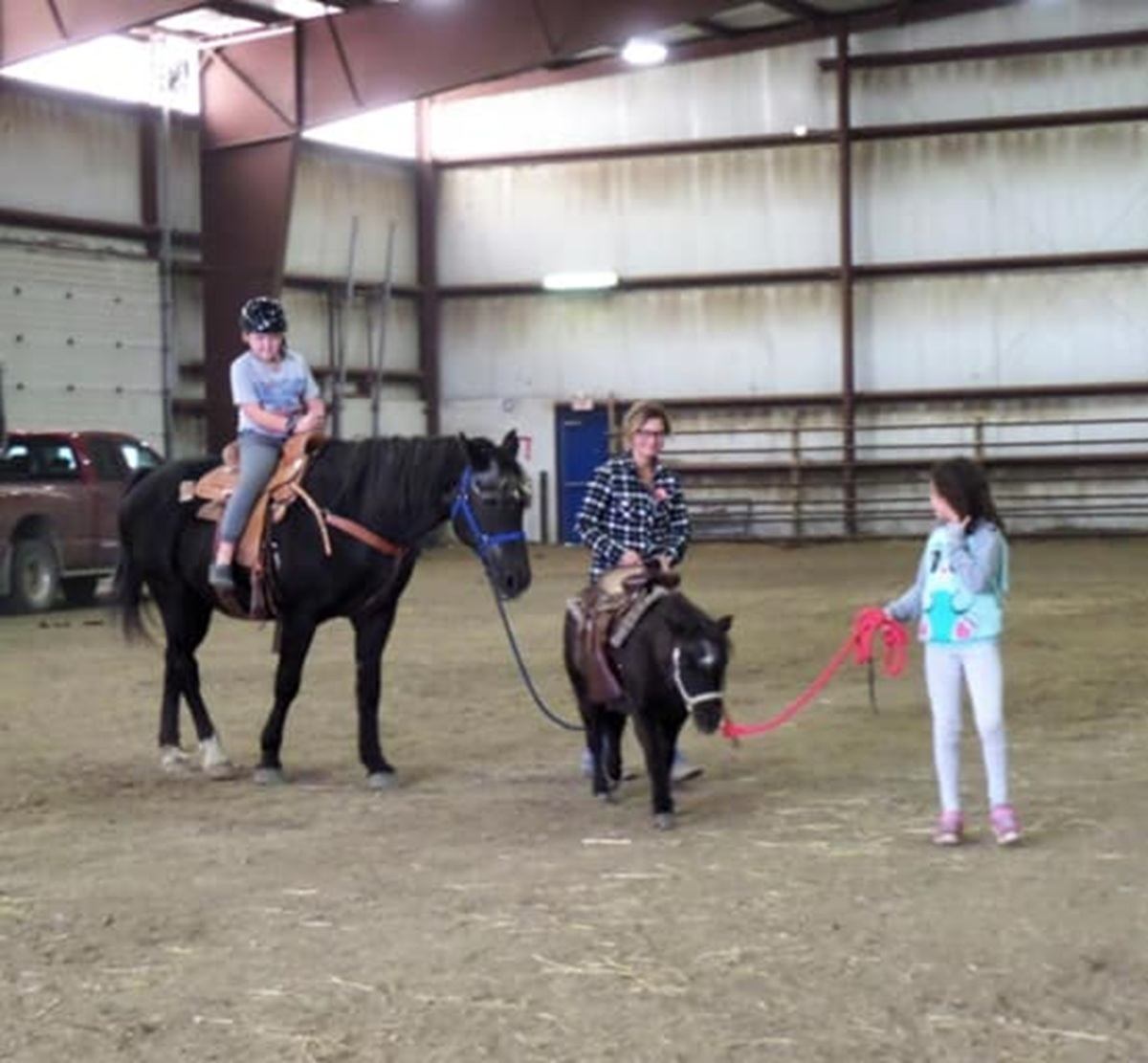 Indoor riding arena with child on horse and two people leading a small pony.