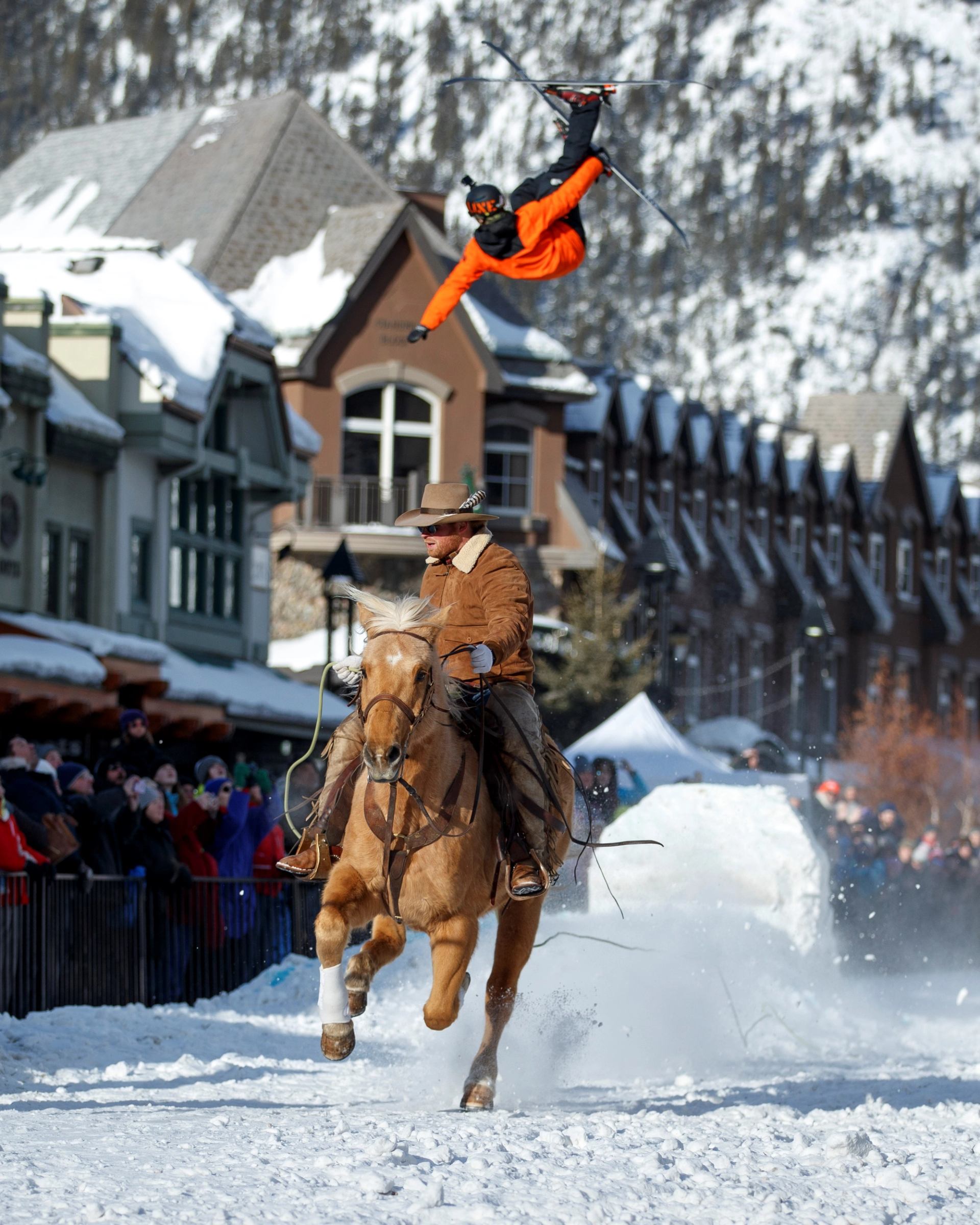 Skier in orange airborne behind galloping horse on snowy street.