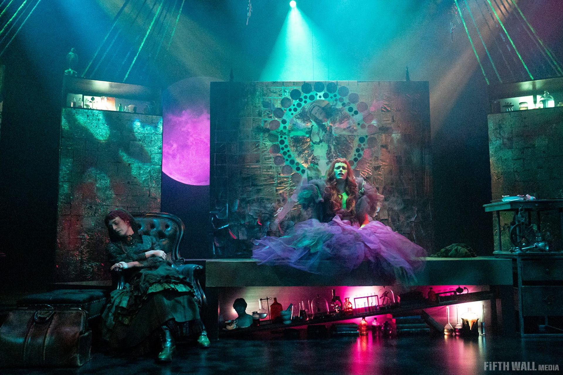 Theatre stage with two performers, purple moon backdrop, and glowing laboratory props.