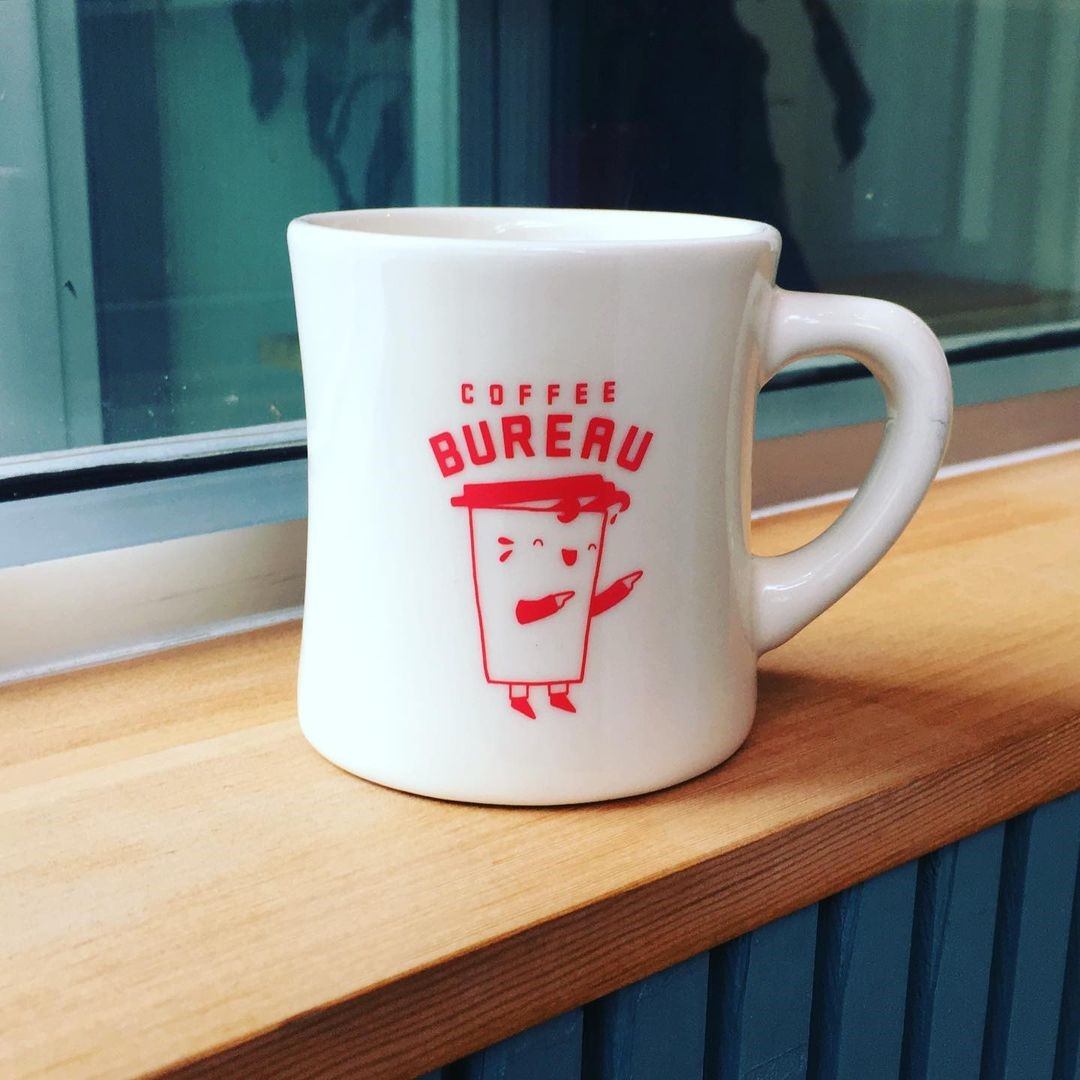 White Coffee Bureau mug with a red cartoon cup logo on a wooden windowsill.