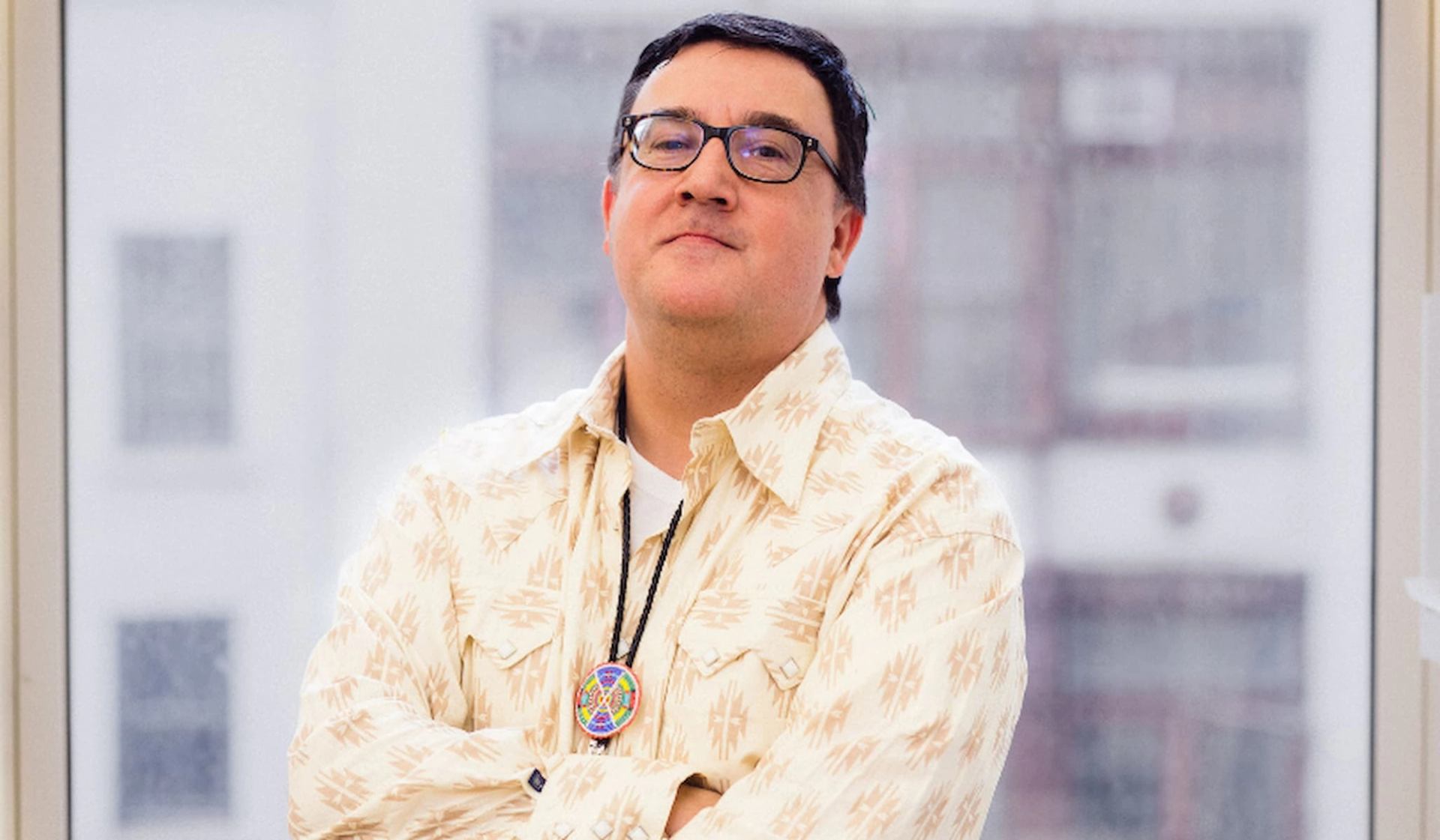 Person wearing patterned shirt and circular pendant stands indoors before large bright windows.